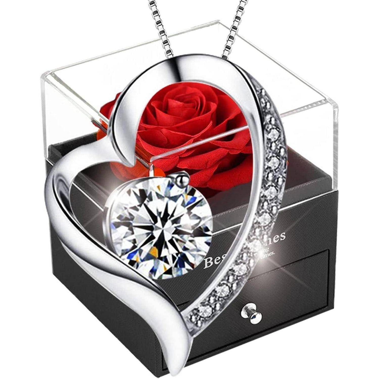 Eine herzförmige silberne Halskette mit einem zentralen Edelstein, besetzt mit kleinen Diamanten, platziert vor einer dekorativen Schatulle mit einer roten Rose.