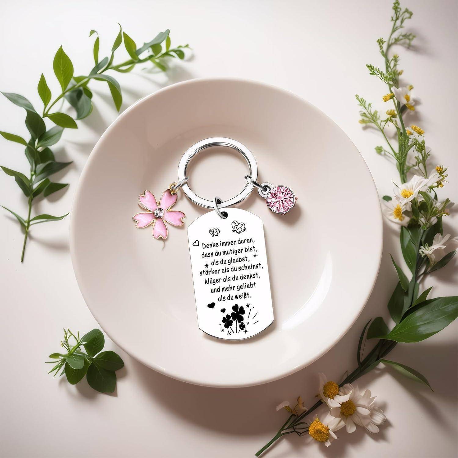 Eine Schlüsselkette mit einer motivierenden Nachricht auf Deutsch, zusammen mit einem pinken Blütenanhänger, liegt auf einem weißen Teller, umgeben von Grünpflanzen und Blumen.