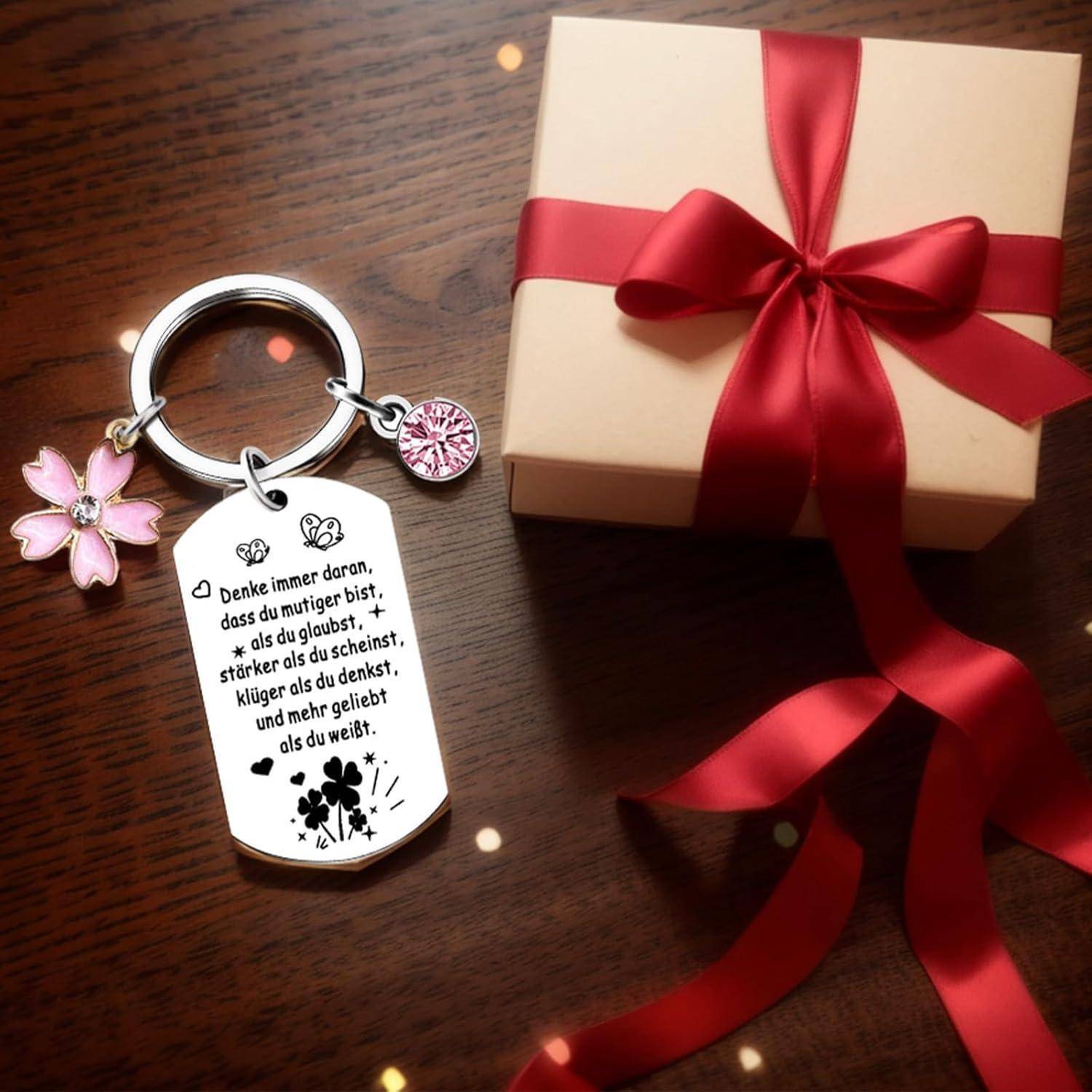 Ein Schlüsselanhänger mit einem Metallanhänger, in Deutsch graviert, zusammen mit einem Kirschblüten-Charm, liegt neben einem eingewickelten Geschenk mit roter Schleife auf einer Holzoberfläche.