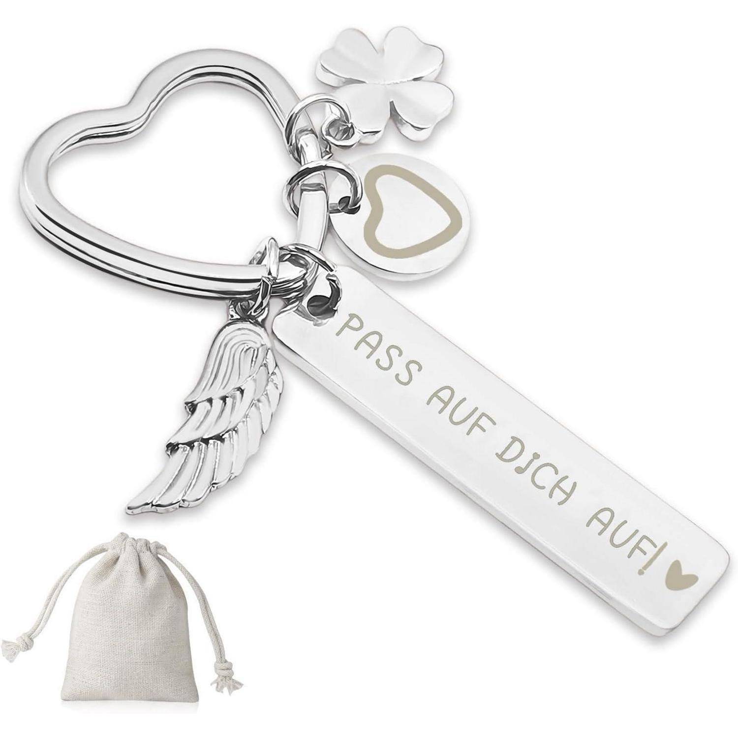 Schlüsselanhänger mit einem Herz, Kleeblatt, Flügel und einem weißen Anhänger mit der Aufschrift 'PASS AUF DICH AUF!'. Eine kleine beige Tasche ist daneben zu sehen.