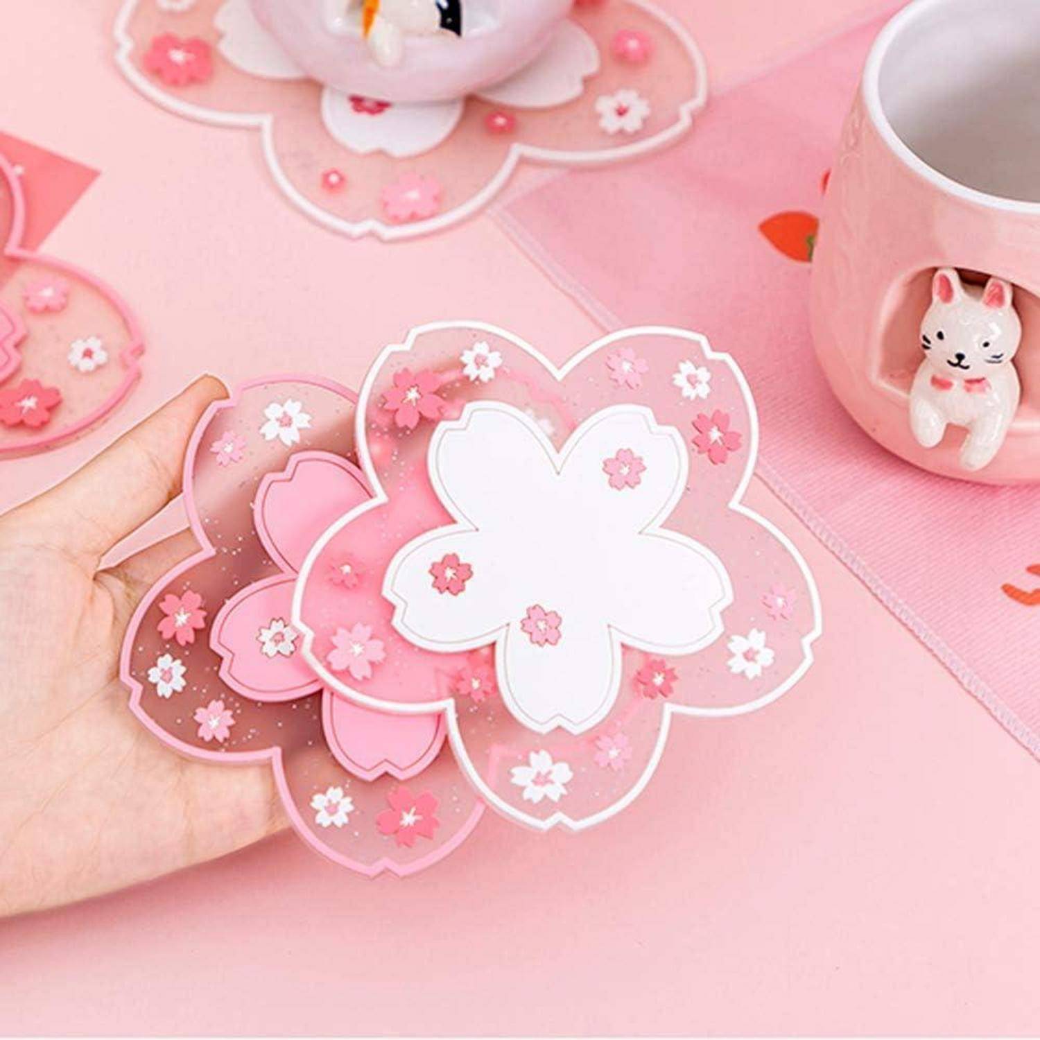 Eine Hand hält rosa kirschblütenförmige Untersetzer in der Nähe einer Tasse mit einer kleinen Katzenfigur am Henkel, die auf einer rosa Oberfläche steht.
