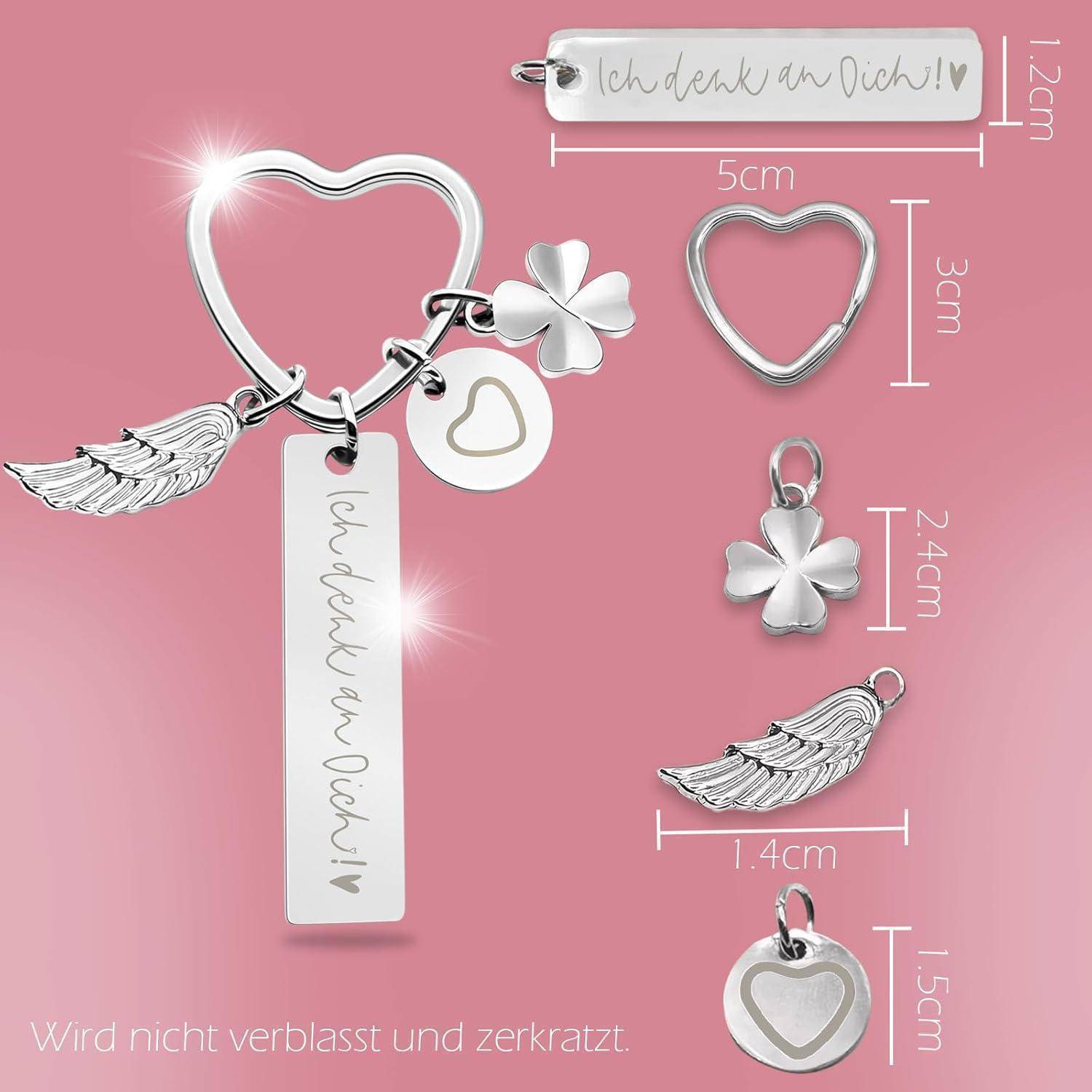 Silbernes Schlüsselanhänger-Set auf rosa Hintergrund mit Herz-, Kleeblatt-, Flügel-Anhängern und einem gravierten Anhänger mit der Aufschrift 