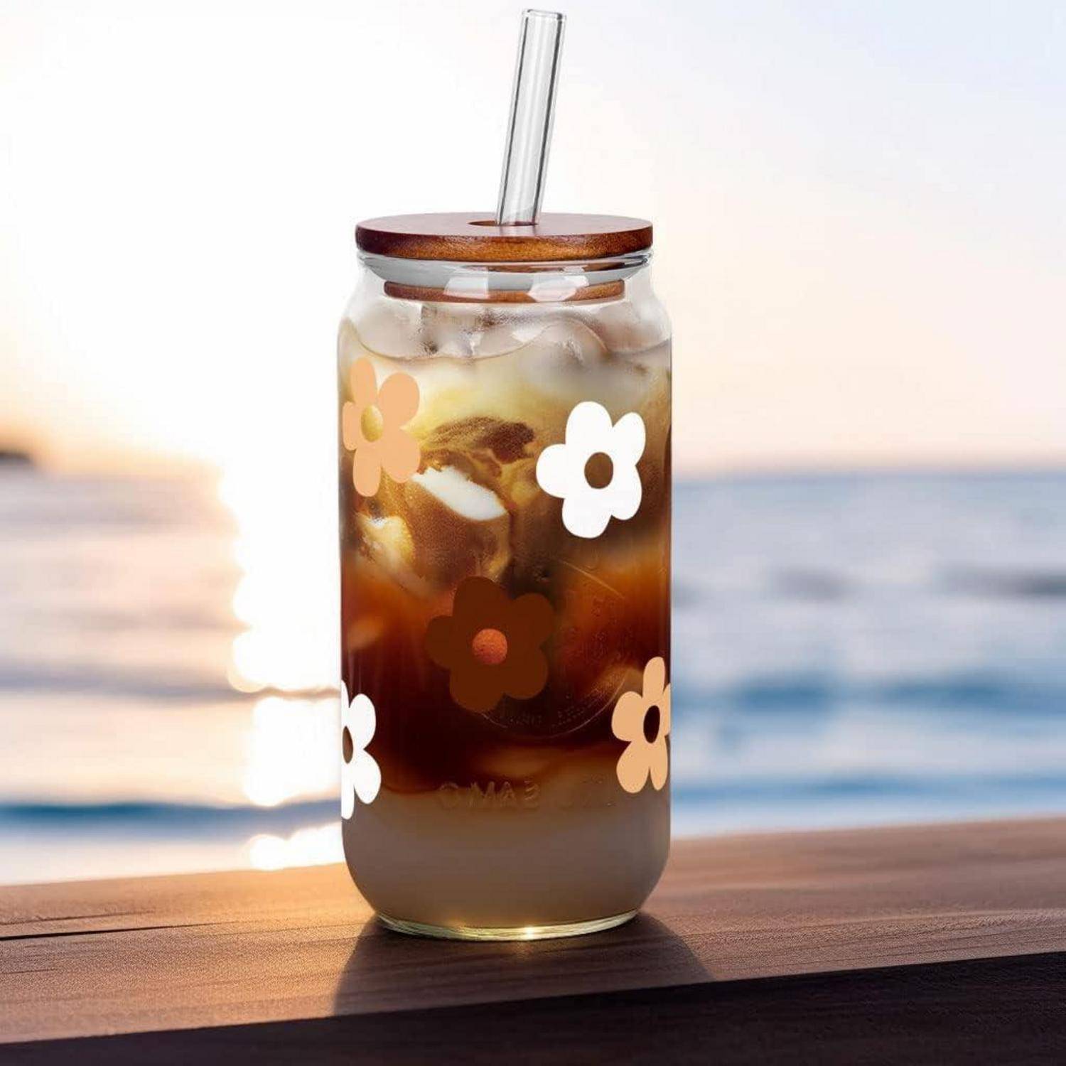 Ein Glas mit Eiskaffee und einem Strohhalm, geschmückt mit Blumenmustern, steht auf einem Holztisch mit Blick auf einen Sonnenuntergang am Strand.