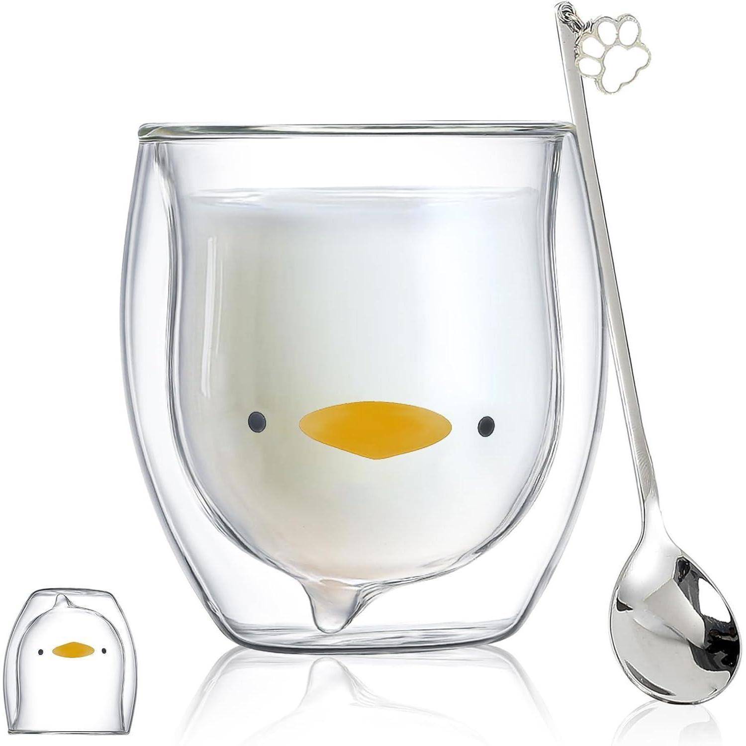 Eine doppelwandige Glasstasse mit einer Pinguingesichtsillustration, die neben einem silbernen Löffel mit einem Pfotenabdruckdesign am Griff präsentiert wird.
