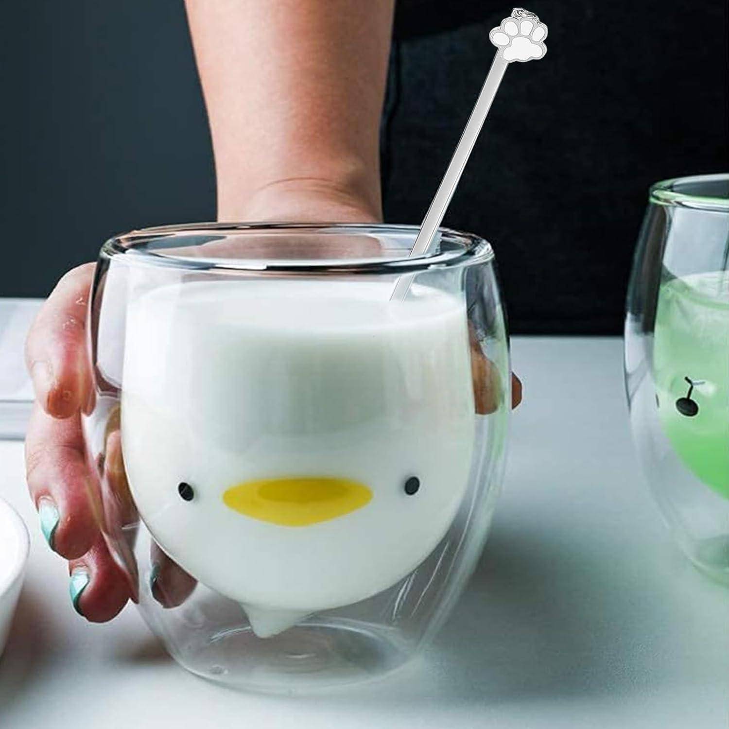 Eine Hand hält ein doppelwandiges Glas mit Milch und zeigt dabei ein Kükendesign. Ein weiteres Glas mit einem anderen Motiv ist teilweise zu sehen.