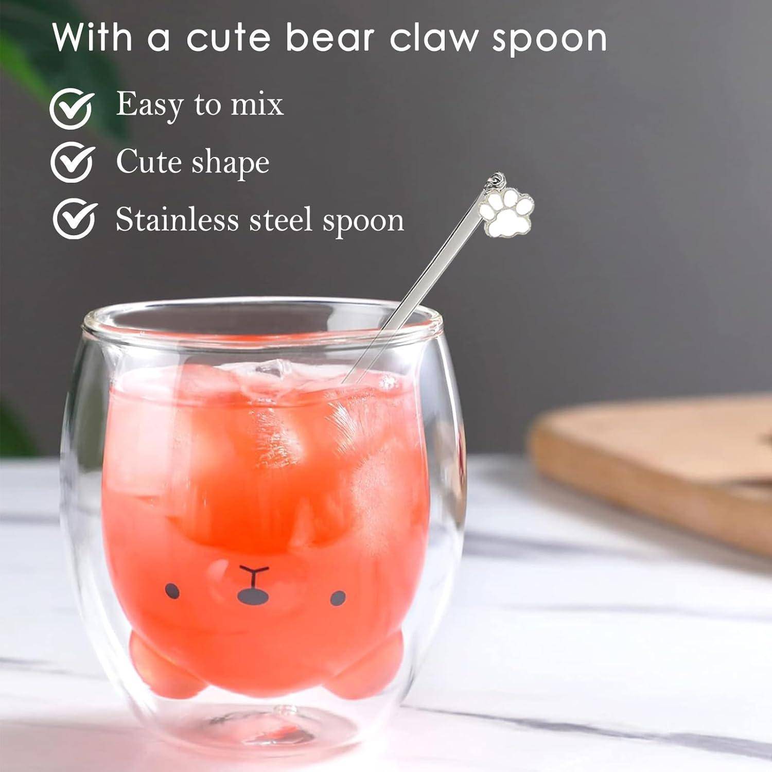 Ein Glasbecher mit einem niedlichen Bärengesicht-Design, gefüllt mit einer orangefarbenen Flüssigkeit. Ein Löffel in Form einer Edelstahlbärentatze befindet sich darin. Der Text lautet: „Leicht zu mischen, Süße Form, Edelstahllöffel
