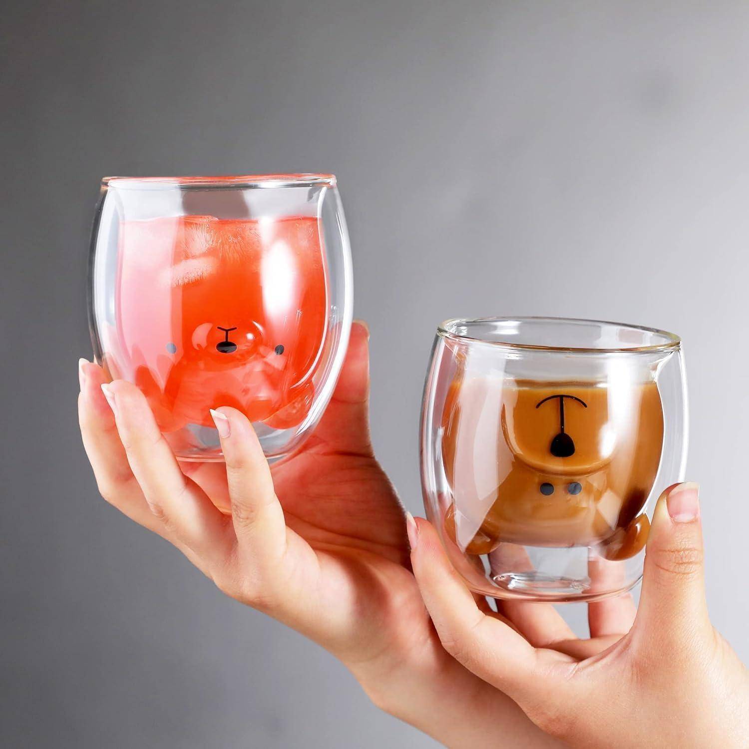 Zwei Hände halten Glasbecher mit Cartoon-Tiermotiven, die orangefarbene und braune Flüssigkeit enthalten. Die Becher sind transparent und doppelwandig.