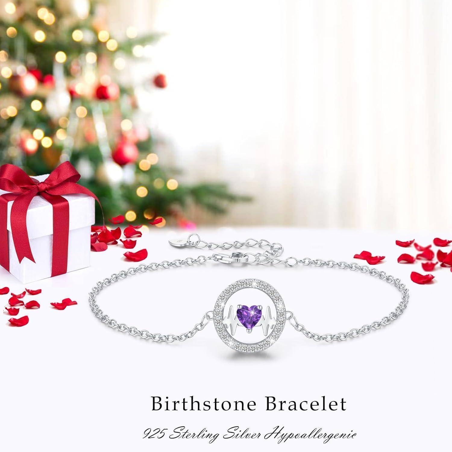 Ein Armband aus Sterlingsilber mit einem herz­förmigen lila Edelstein in der Mitte. Links davon eine Geschenkbox und verstreute Blütenblätter, mit einem unscharfen Weihnachtsbaum im Hintergrund. Text lautet: 'Geburtsstein-Armband'.