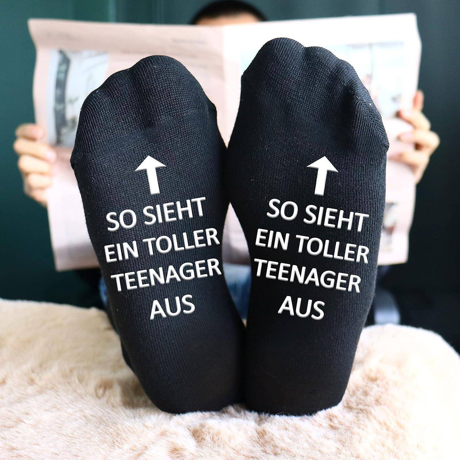 Eine Person entspannt sich mit hochgelegten Füßen, trägt schwarze Socken mit dem Text ‚SO SIEHT EIN TOLLER TEENAGER AUS'. Die Person hält eine Zeitung.