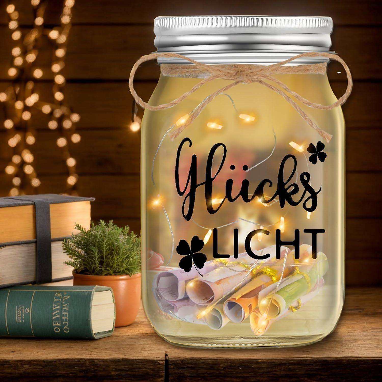 Ein Glasgefäß gefüllt mit warmen Lichterketten und bunten aufgerollten Papierstreifen, beschriftet mit 'Glücks Licht', steht auf einem Holztisch mit Büchern und einer Pflanze.