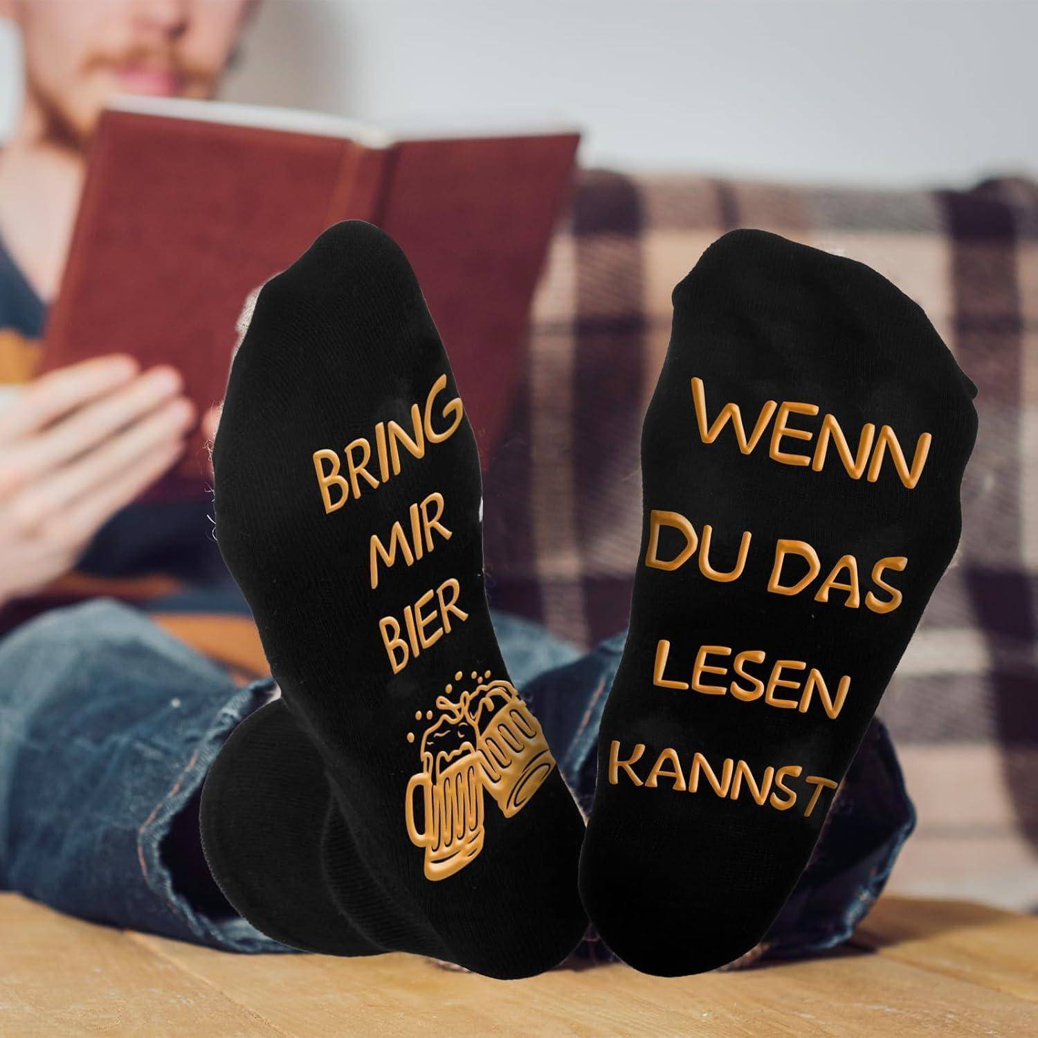 Person liest ein Buch auf einer Couch, Füße in schwarzen Socken mit dem Text: 'Bring mir Bier, wenn du das lesen kannst'.