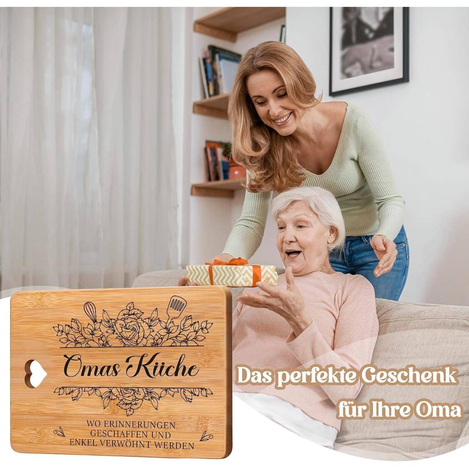 Eine Frau überreicht einer älteren Dame ein Geschenk mit einem Lächeln. Der Text auf dem Schneidebrett lautet „Omas Küche