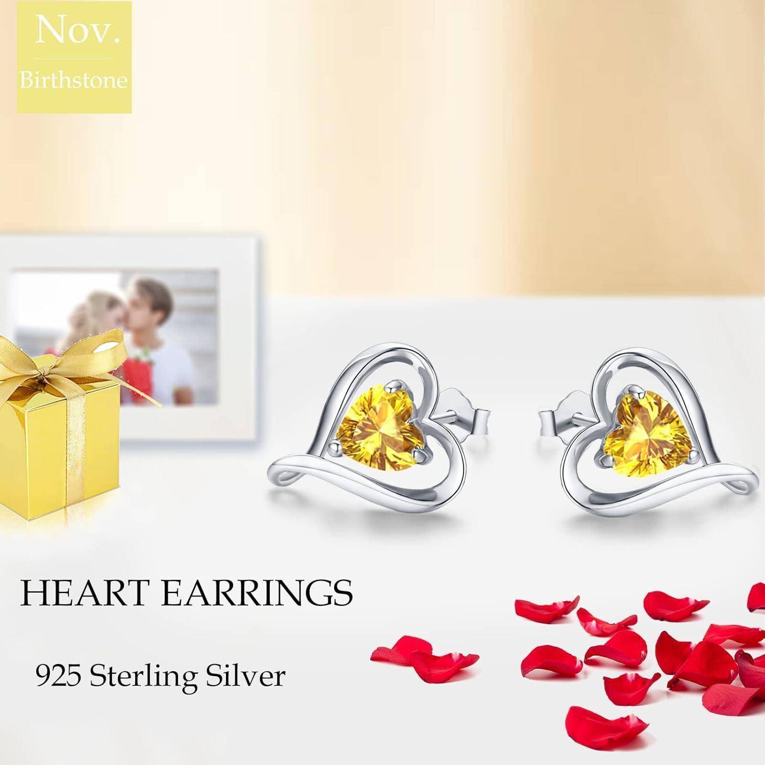 Herzförmige Ohrringe mit gelben Steinen neben Rosenblütenblättern, einer Geschenkbox und einem unscharfen Familienfoto; beschriftet mit '925 Sterling Silber'.