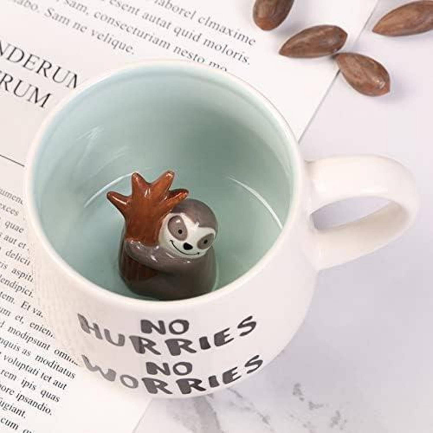 Eine weiße Tasse mit dem Schriftzug 'Keine Eile, keine Sorgen', mit einer Faultier-Figur im Inneren. Die Tasse steht neben Mandeln und Papier auf einem Tisch.
