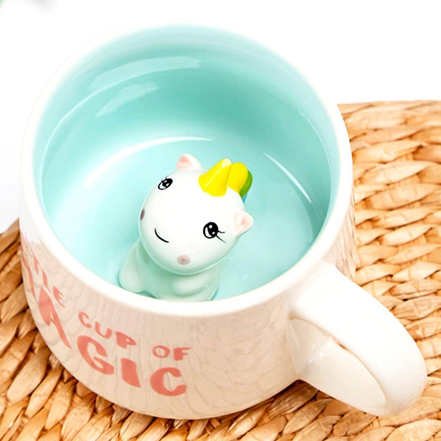Eine Tasse mit den Worten 'Eine Tasse Magie' enthält eine kleine Einhornfigur im Inneren, umgeben von einer hellblauen Innenfläche, auf einer gewebten Matte.