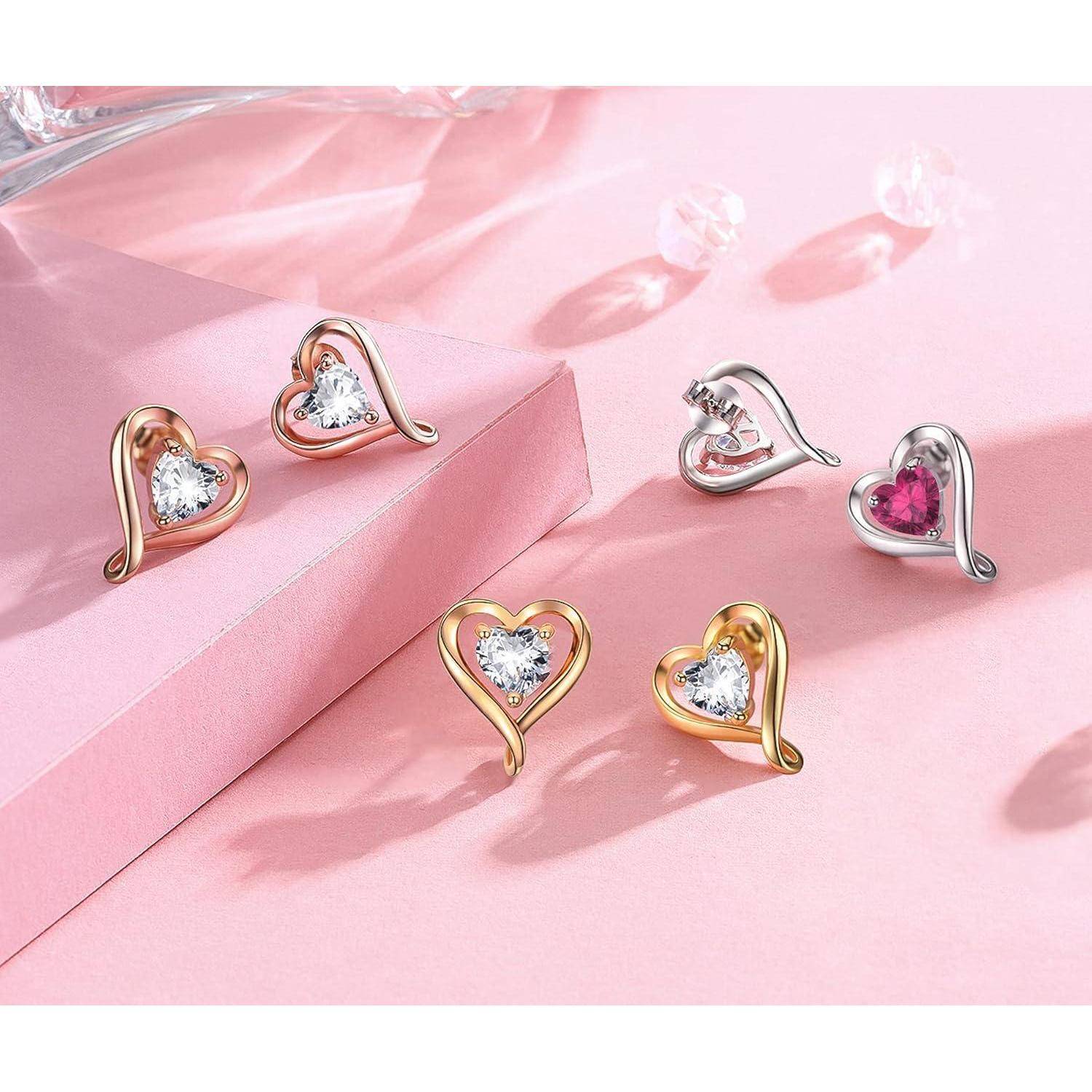 Herzförmige Ohrringe mit Edelsteinen, die auf einer rosa Oberfläche ausgestellt sind. Ein Paar ist golden mit klaren Steinen, und das andere ist silbern mit einem rosafarbenen Stein.