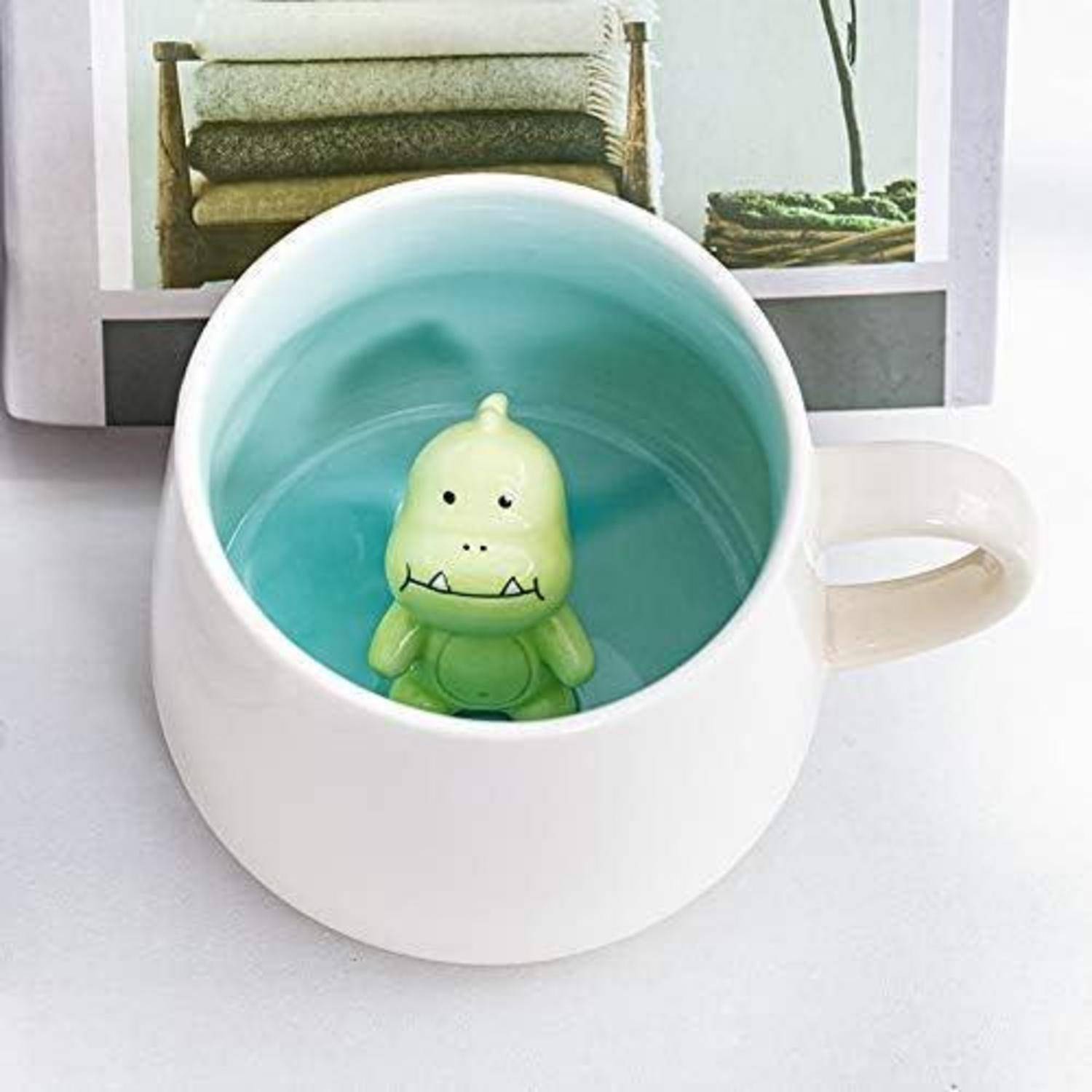 Eine weiße Tasse, gefüllt mit einer hellblauen Flüssigkeit, enthält einen kleinen grünen Spielzeugdinosaurier am Boden. Im Hintergrund befinden sich ein Stapel zusammengelegter Handtücher und eine Pflanze.