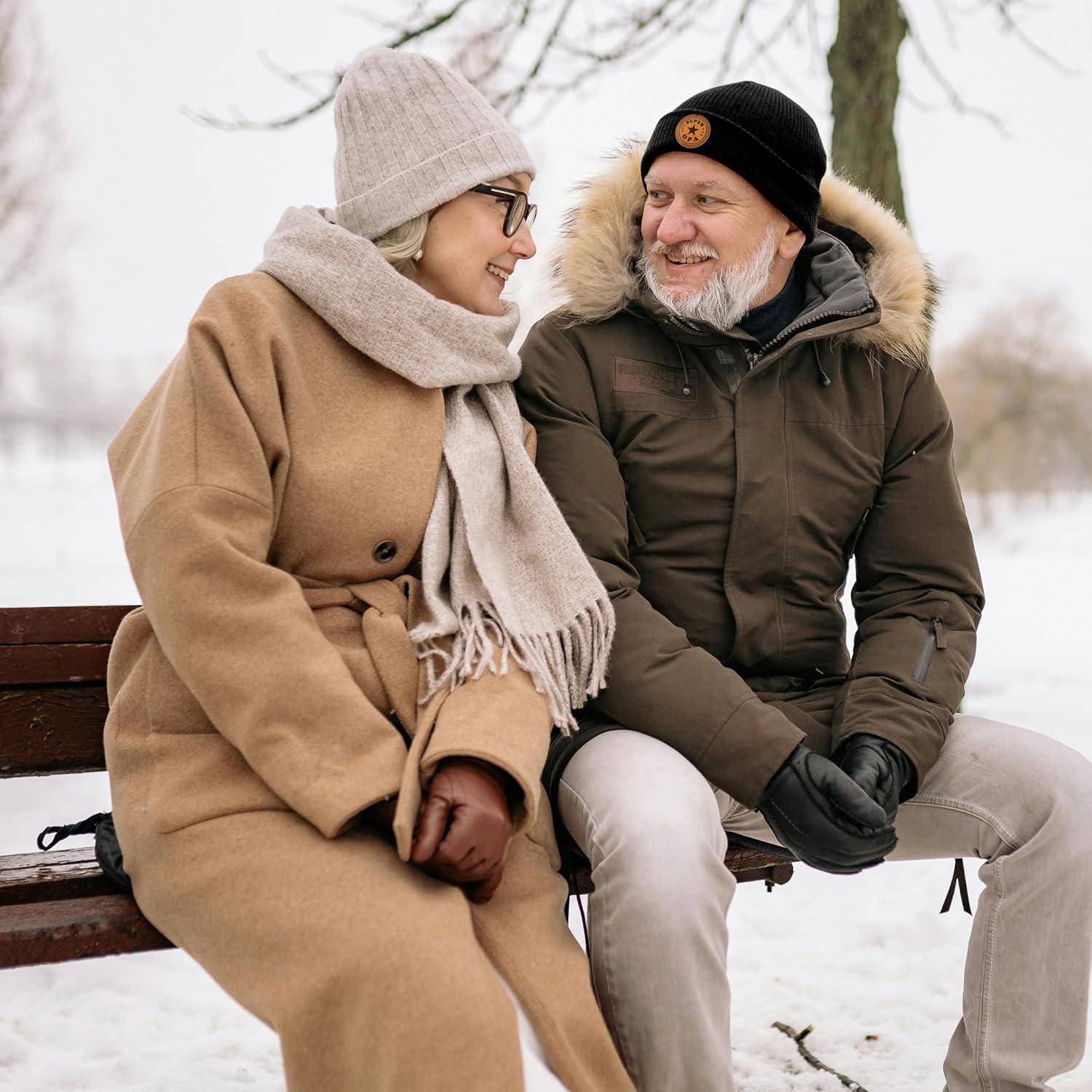 Ein älteres Paar in Winterkleidung sitzt auf einer Parkbank, lächelt und unterhält sich. Schneebedeckte Bäume befinden sich im Hintergrund.