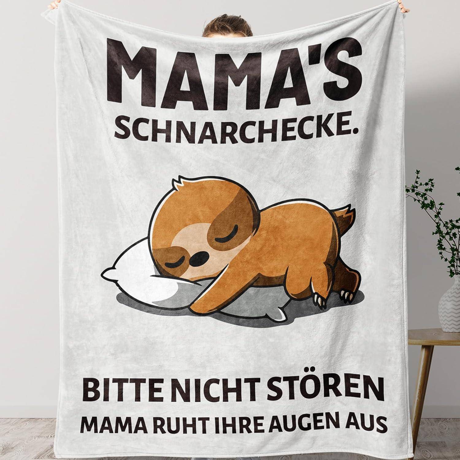 Eine Person hält eine Decke mit einem Cartoon-Faultier, das auf einem Kissen schläft. Text lautet: 'Mamas Schnarchecke. Bitte nicht stören. Mama ruht ihre Augen aus.