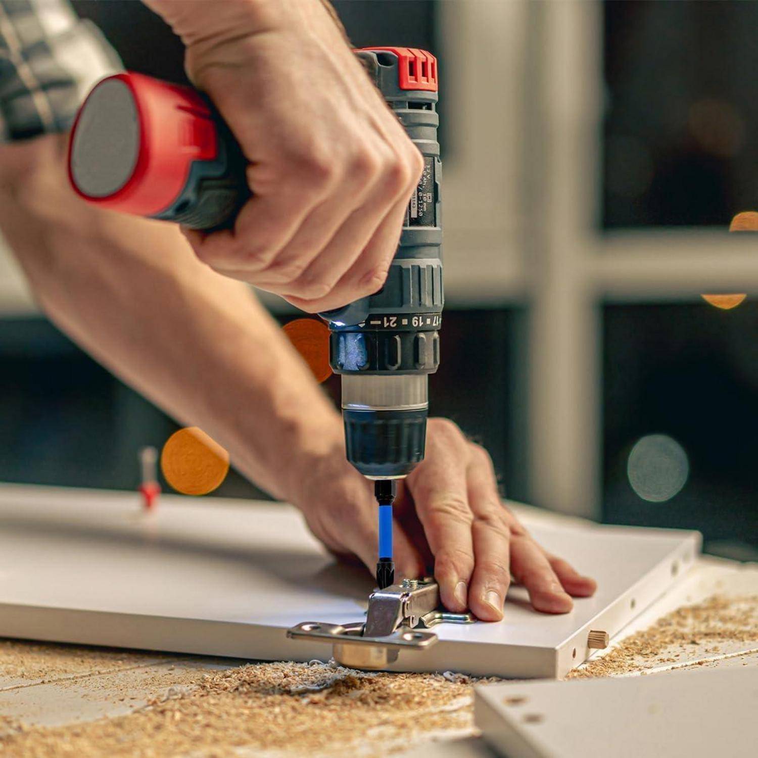 Eine Person benutzt eine Bohrmaschine an einem Scharnier, das an einer Holzplatte befestigt ist und deutet damit auf laufende Montage- oder Reparaturarbeiten hin.
