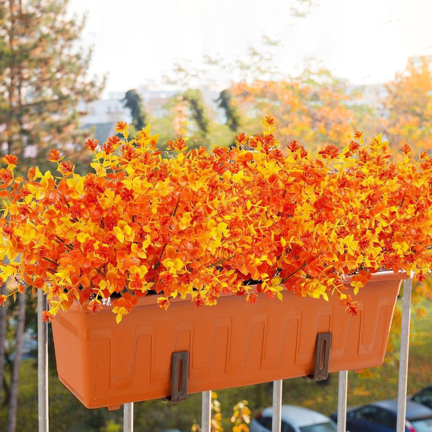 Ein heller orangefarbener Blumenkasten, gefüllt mit lebhaft gelb-orangefarbenen Blumen, ist an einem Balkongeländer angebracht und vor einer unscharfen Baumkulisse platziert.