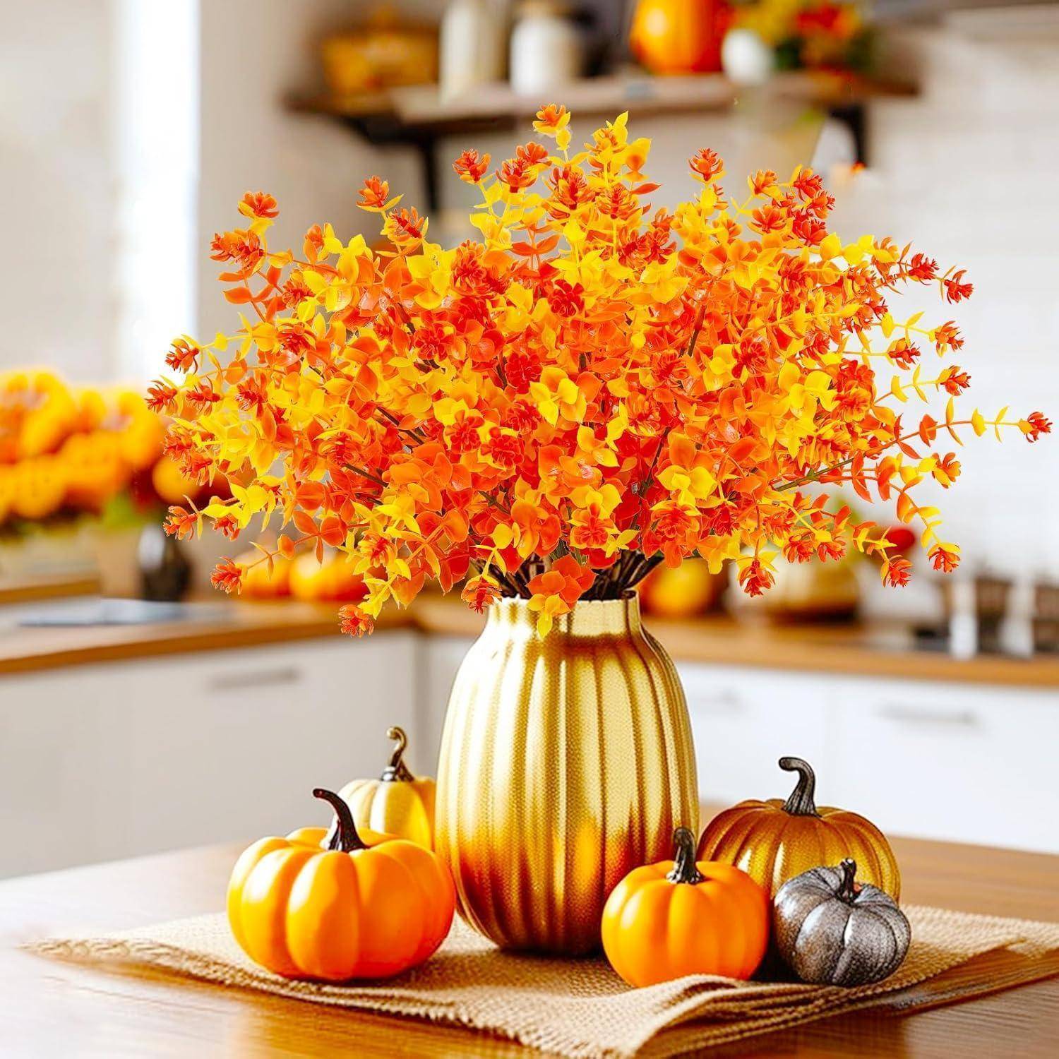 Eine goldene Vase enthält lebendige orangefarbene Herbstblätter, umgeben von kleinen Kürbissen auf einem Holztisch, was eine warme Herbst-Kücheneinrichtung schafft.