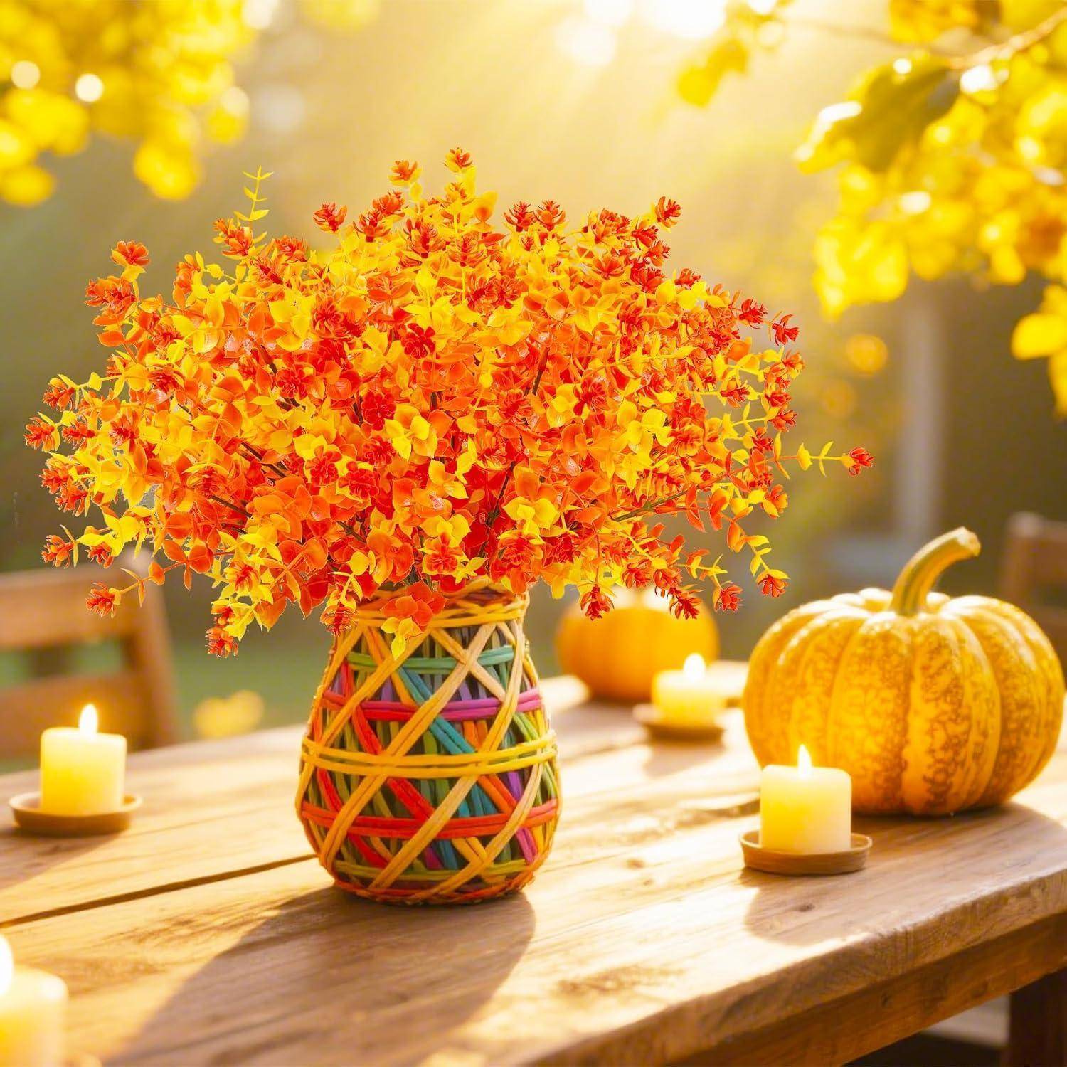 Eine farbenprächtige Vase, gefüllt mit leuchtend orangen und gelben Herbstblättern auf einem Holztisch im Freien. Um die Vase herum brennende Kerzen und ein kleiner Kürbis, während Sonnenlicht die Szene erhellt.