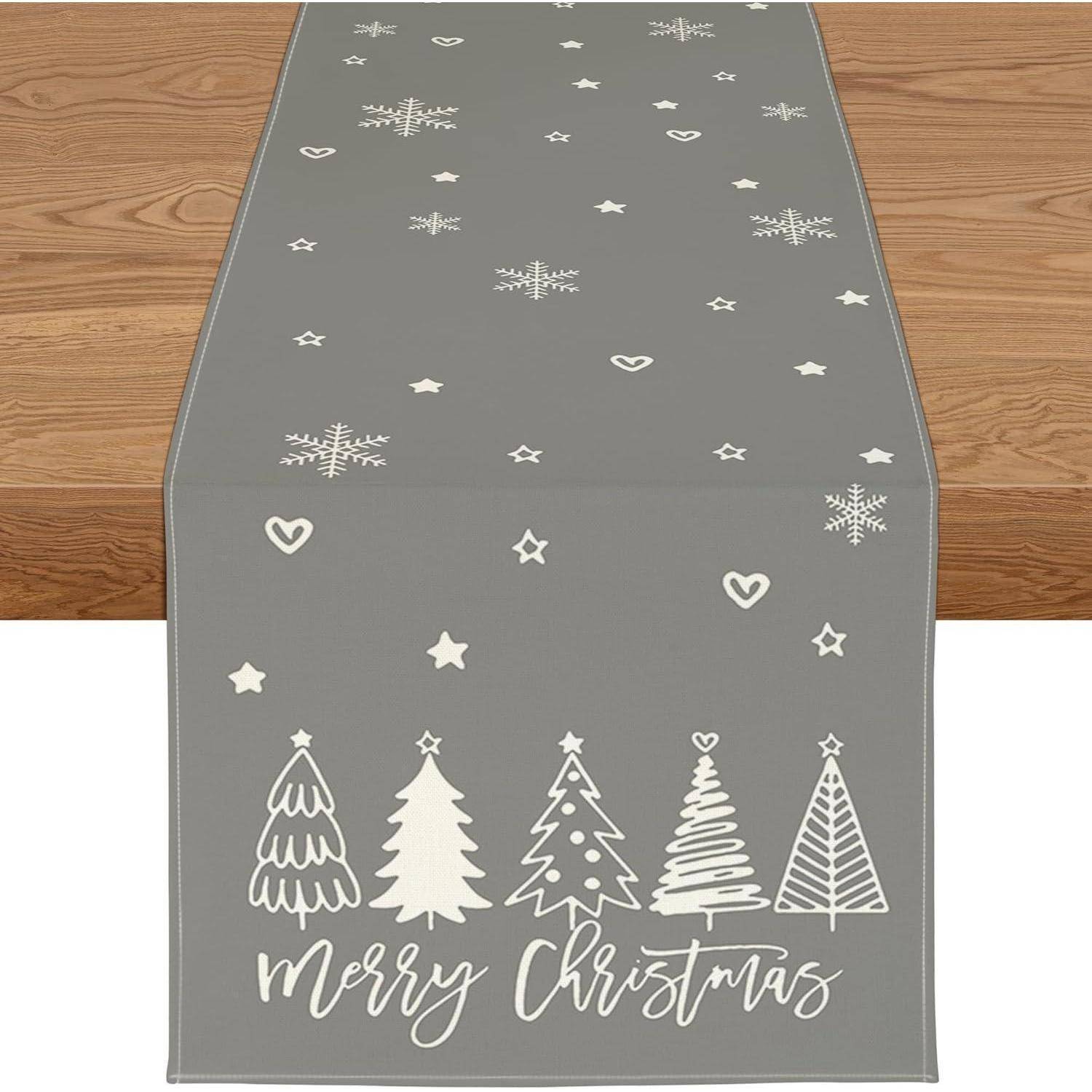Grauer Tischläufer mit 'Frohe Weihnachten'-Schriftzug und weißen Umrissen von Weihnachtsbäumen, Sternen und Schneeflocken, der über einem Holztisch drapiert ist.