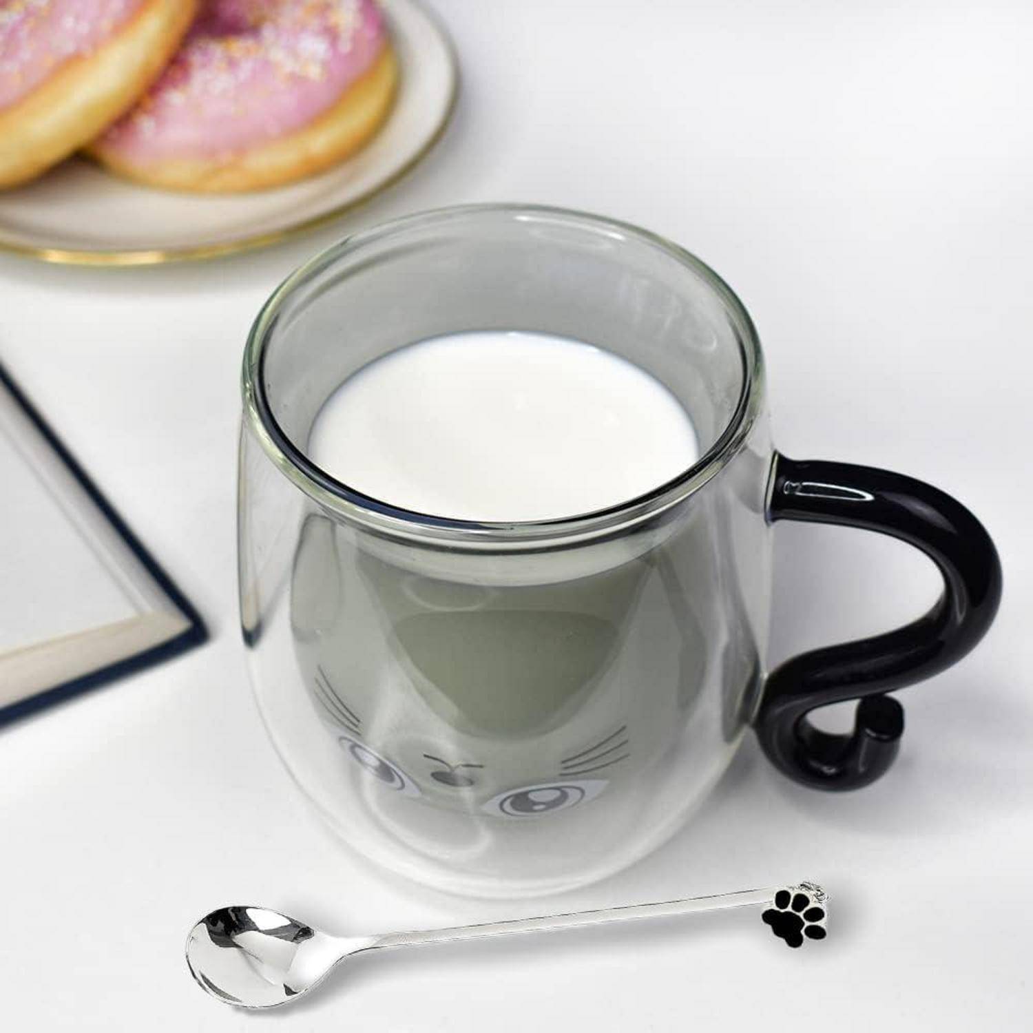 Eine Tasse mit einer Katzengesichtdesign, gefüllt mit Milch, begleitet von einem Löffel mit einem Pfotenabdruck und einem Teller mit Donuts im Hintergrund.