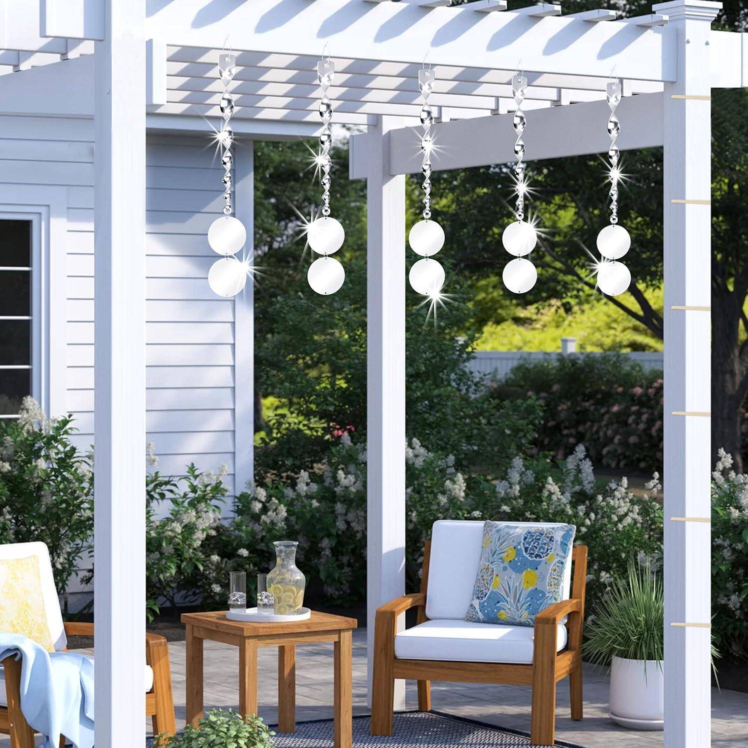 Ein weißer Pergola mit hängenden Kugelleuchten, zwei Holzstühlen mit Blumenkissen, einem kleinen Tisch und Grünpflanzen im Hintergrund.