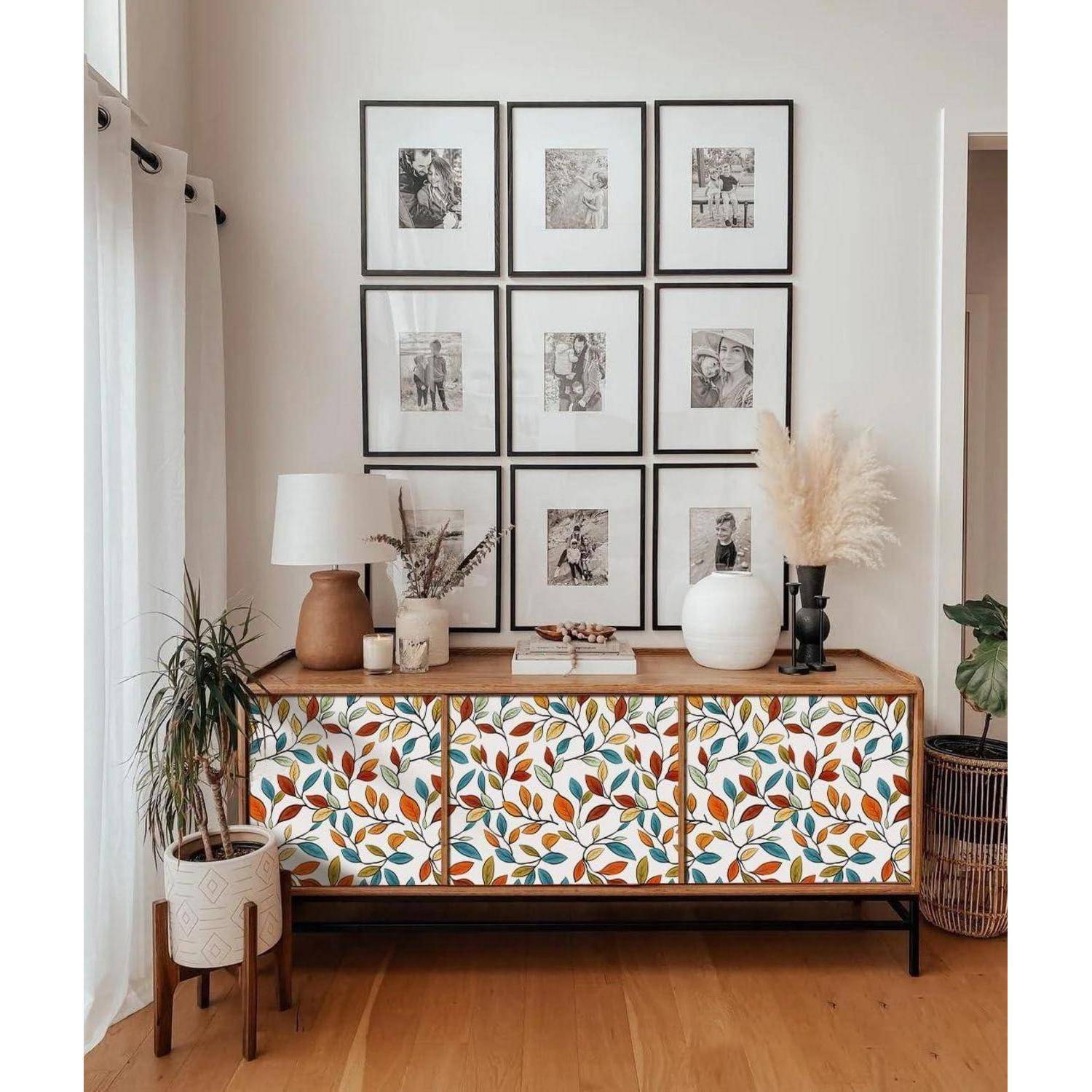 Ein dekoriertes Sideboard mit bunten Blattmustern an den Schranktüren, bekrönt von Vasen, Büchern und einer Lampe. Darüber ein Raster aus gerahmten Fotos.