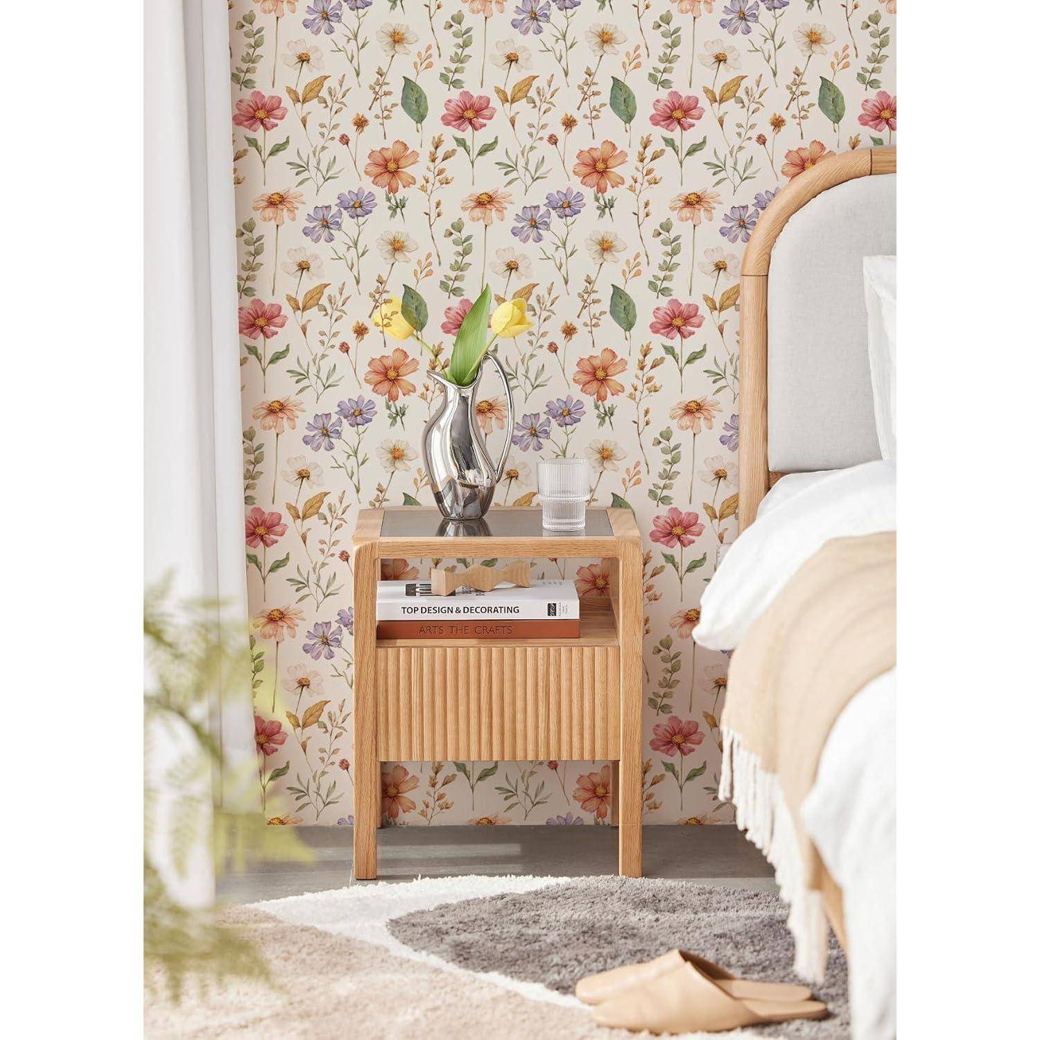 Nachttisch mit einer silbernen Vase, in der Tulpen stehen, und einem Glas Wasser, neben einem Bett mit beige-weißer Bettwäsche, vor einer Blumentapete.