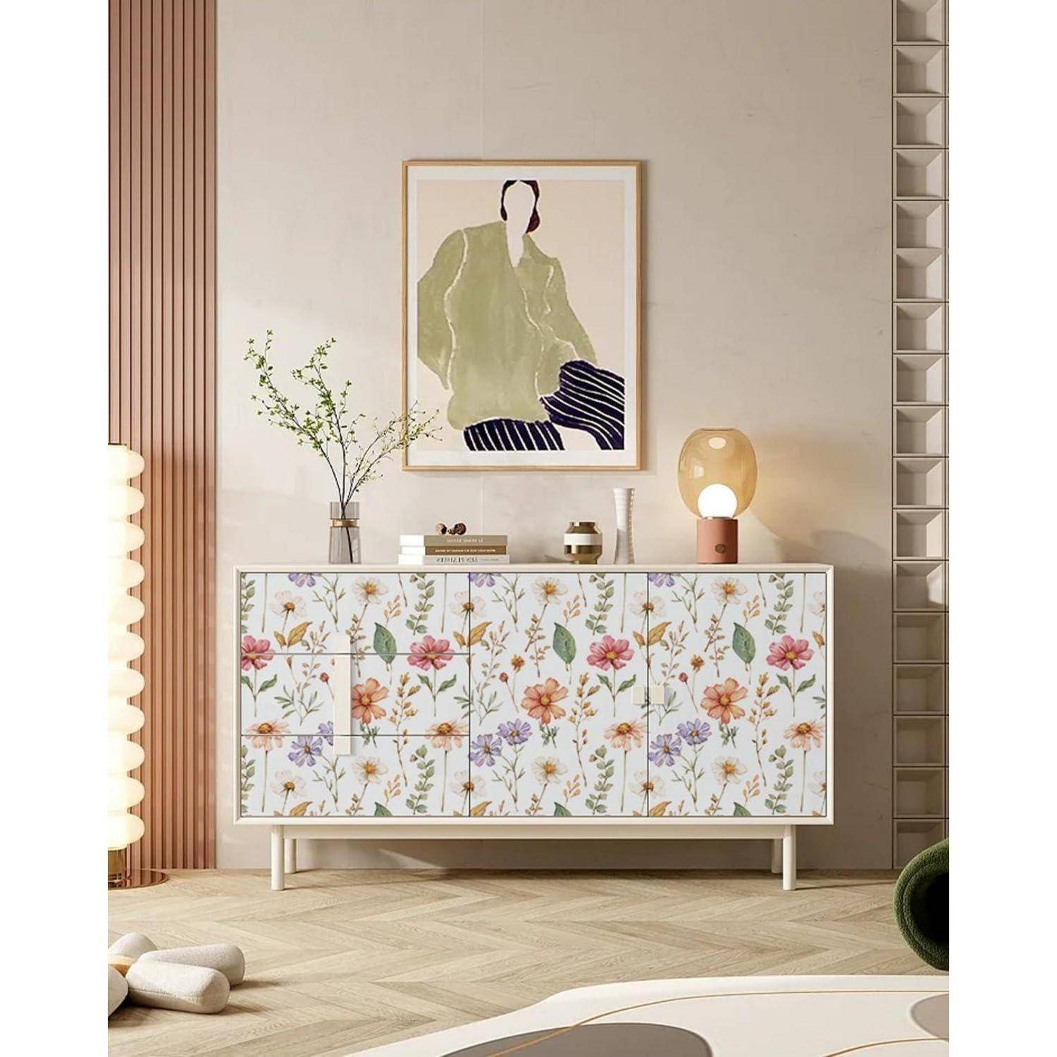 Ein minimalistisches Wohnzimmer zeigt ein Sideboard mit Blumenmuster, ein gerahmtes abstraktes Kunstwerk darüber, eine Vase mit Zweigen und eine kleine Tischlampe.