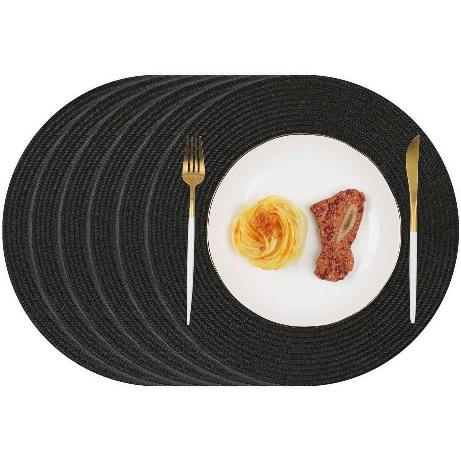 Ein Teller mit Pasta und einem Stück Fleisch ist auf einer großen schwarzen ovalen Tischunterlage zentriert. Eine Gabel und ein Messer sind rechts und links davon platziert.