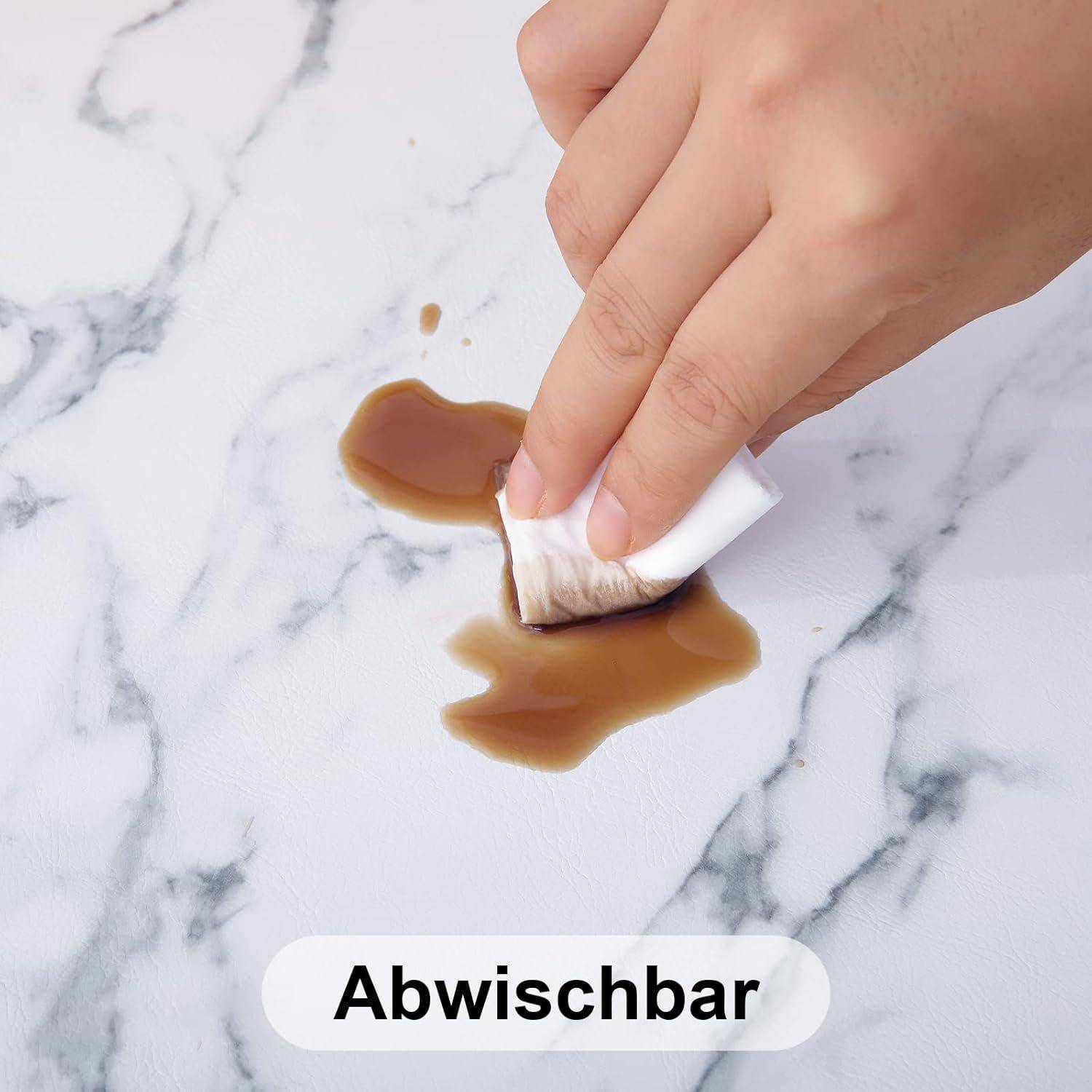 Eine Hand, die einen Flüssigkeitsfleck auf einer Marmeroberfläche mit einem Tuch abwischt. Text darunter liest sich 'Abwischbar', was anzeigt, dass die Oberfläche abwischbar ist.