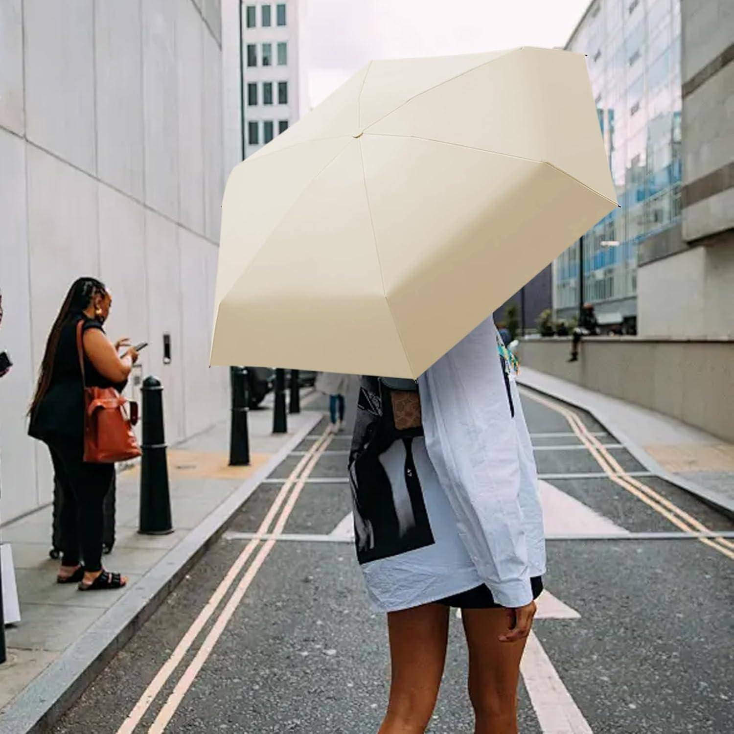 Eine Person geht an einem regnerischen Tag mit einem großen beigen Regenschirm durch die Stadt, wobei Gebäude und einige Fußgänger im Hintergrund zu sehen sind.