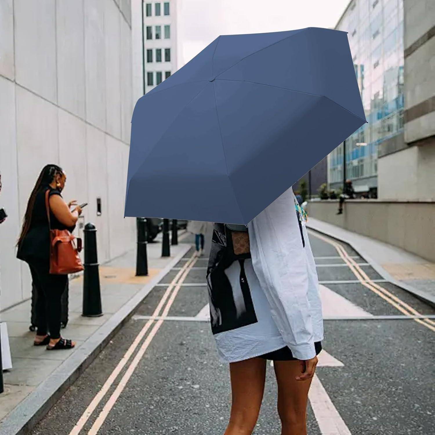 Eine Person hält einen großen marineblauen Regenschirm und geht eine städtische Straße entlang. Im Hintergrund sind weitere Personen in der Nähe eines Gebäudes zu sehen.