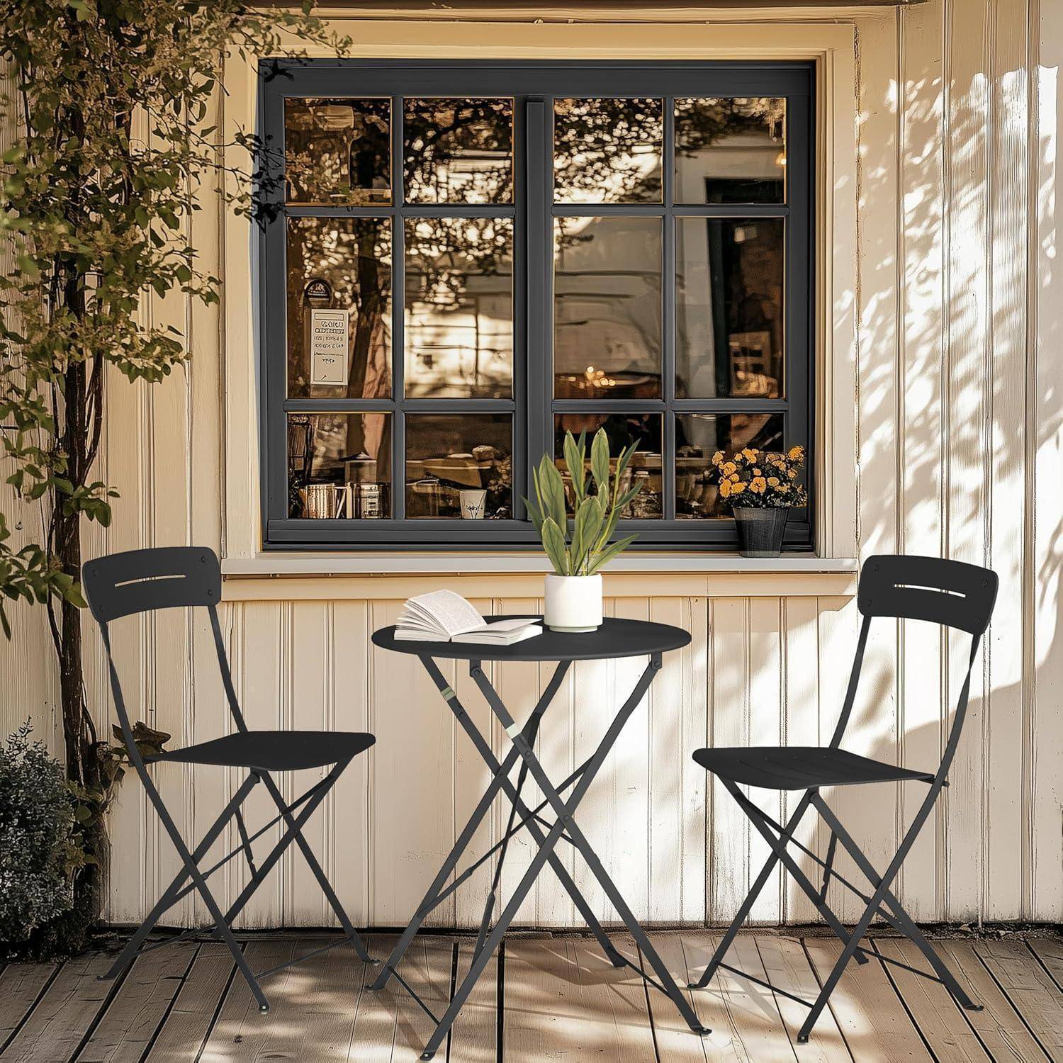 Zwei schwarze Stühle und ein Tisch mit einer Topfpflanze und einem aufgeschlagenen Buch befinden sich auf einer Holzterrasse vor einem Fenster, das nahegelegene Bäume spiegelt.