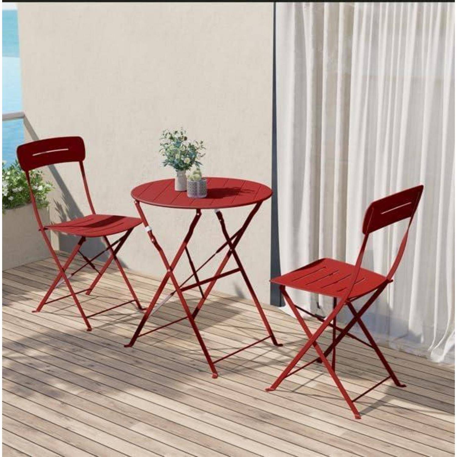 Rotes Outdoor-Bistro-Set mit zwei Stühlen und einem Tisch auf einer Holzterrasse, neben einem Vorhang und Pflanzen; Ozean im Hintergrund.