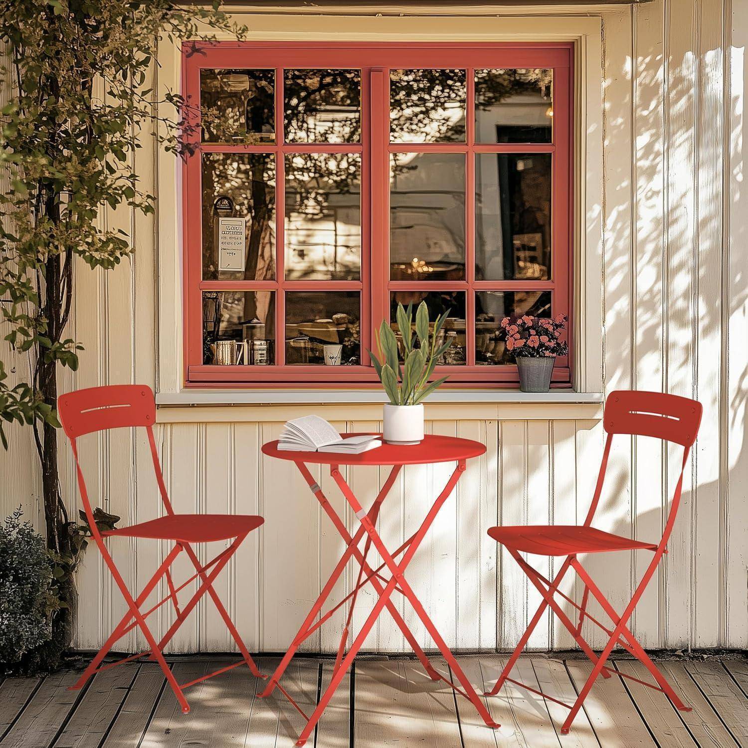 Ein roter Tisch und zwei Stühle stehen auf einer Holzterrasse vor einem cremefarbenen Haus mit einem Fenster. Eine Pflanze und ein Buch liegen auf dem Tisch.