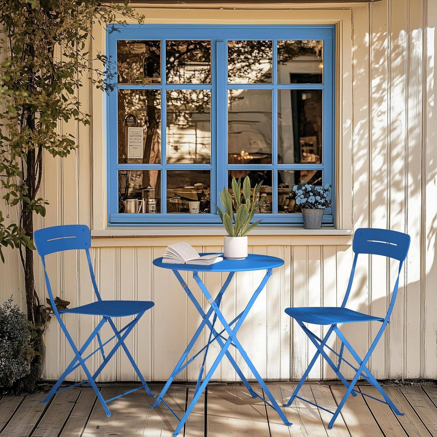 Eine kleine Terasse zeigt einen blauen Metalltisch mit einer Pflanze und einem aufgeschlagenen Buch, flankiert von zwei blauen Stühlen, vor einem Fenster.
