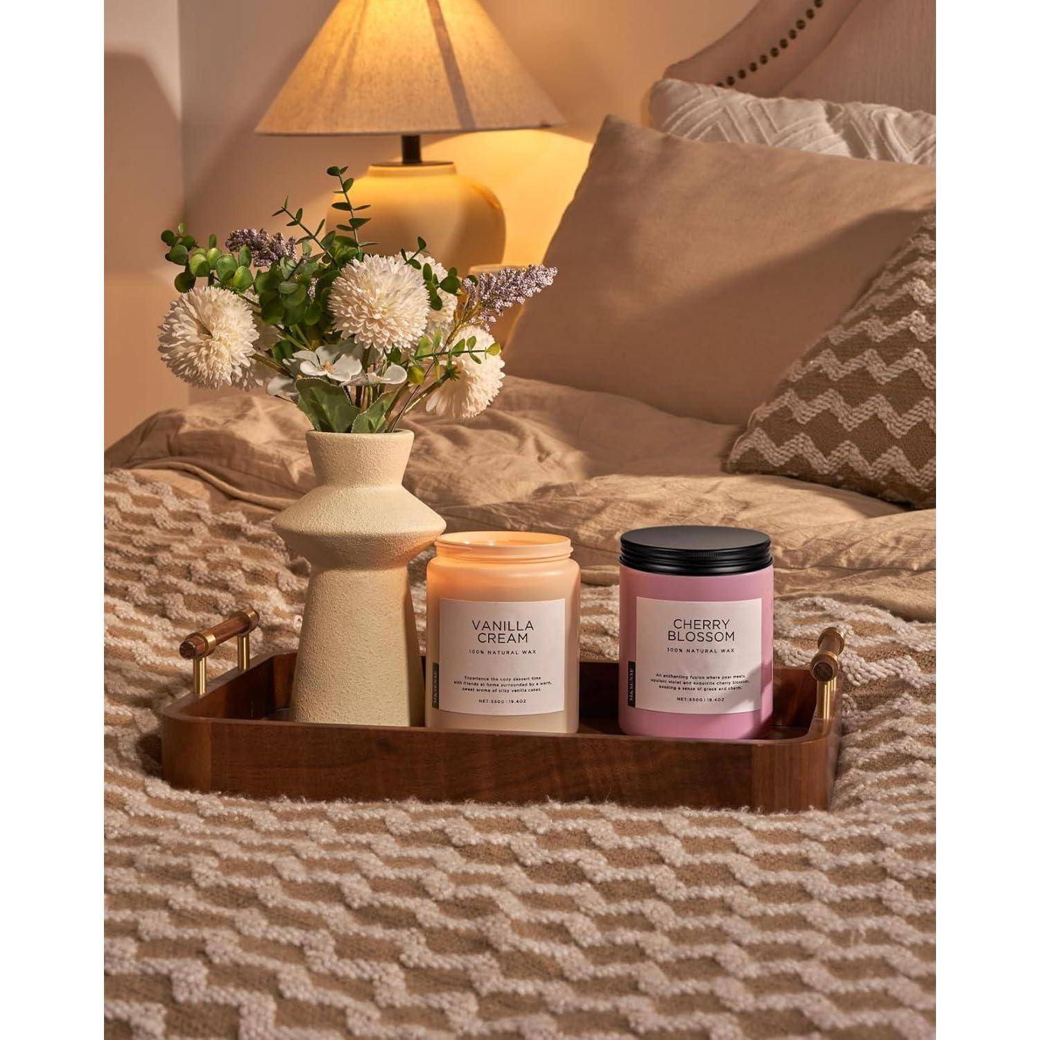 Ein gemütliches Schlafzimmer mit einem Holztablett auf dem Bett. Das Tablett trägt eine Vase mit frischen Blumen und zwei duftenden Kerzen mit den Bezeichnungen ‚Vanillecreme' und ‚Kirschblüte'.