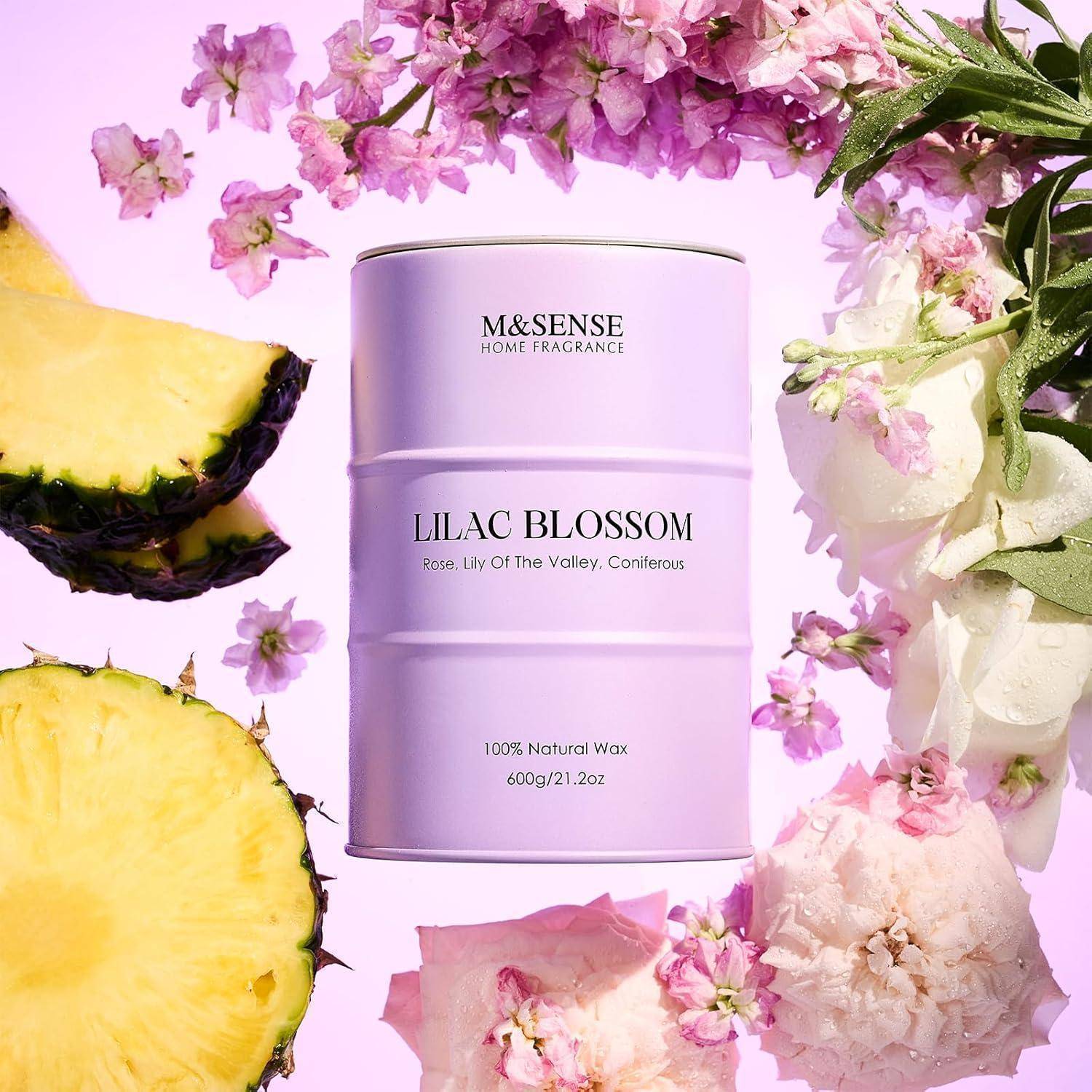 Eine lavendelfarben Kerze mit der Aufschrift ‚Fliederblüte', umgeben von tropischen Früchten und rosa Blumen, vor einem lebhaften Hintergrund.