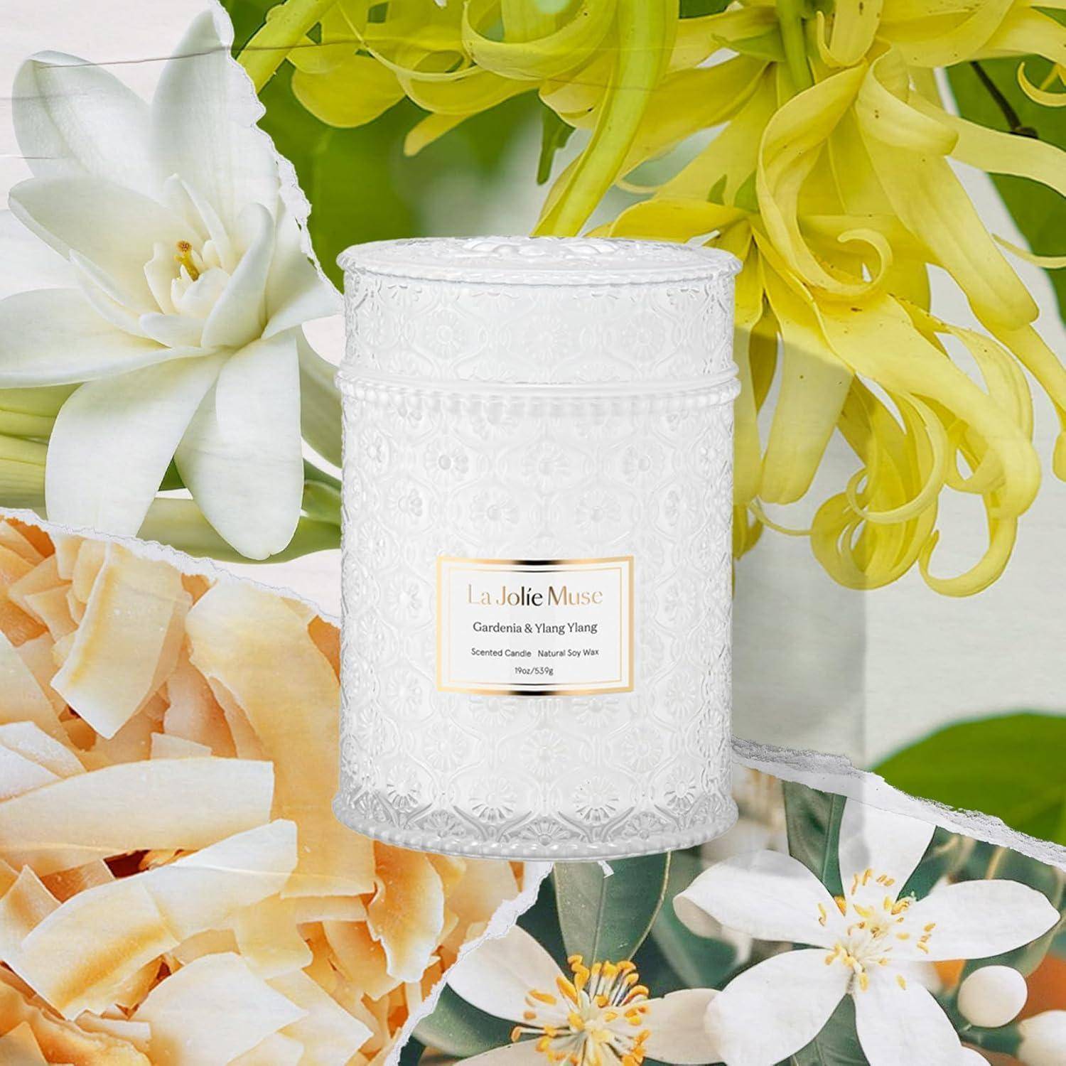 Eine weiße Dekokerze mit der Aufschrift 'La Jolie Muse', umgeben von weißen und gelben Blumen, darunter Gardenien und Ylang-Ylang-Blüten.