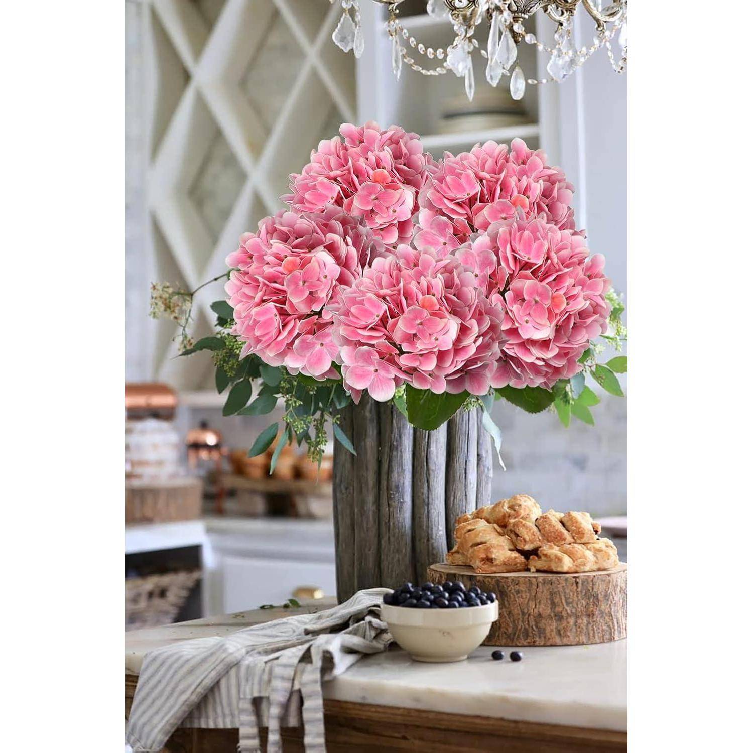 Eine Holzvase mit lebhaft pinken Hortensien steht auf einer Küchenarbeitsfläche, begleitet von Gebäck und einer kleinen Schüssel mit Blaubeeren, unter einem Kronleuchter.