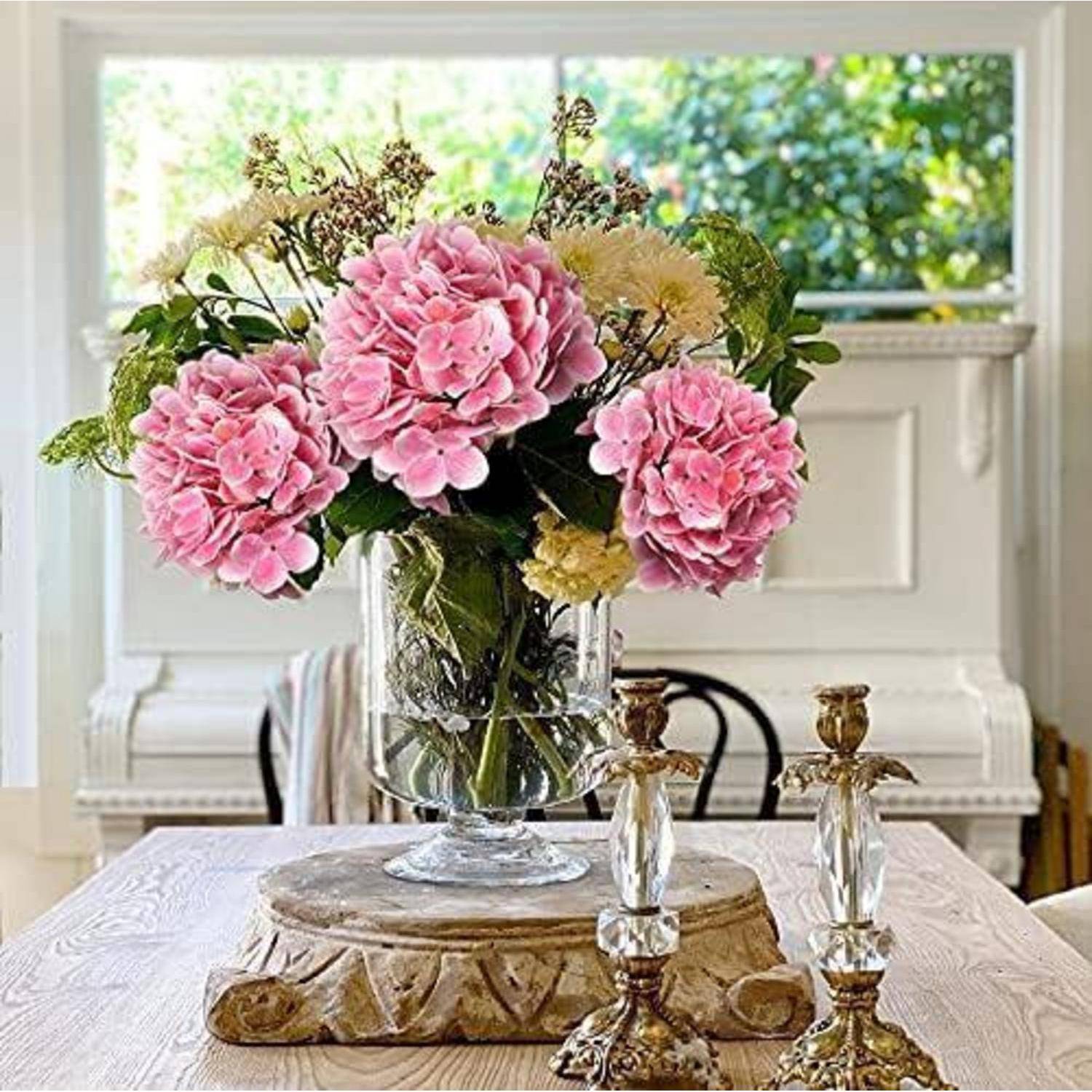 Eine Vase mit rosa Hortensien und gemischten Blumen auf einem Holztisch mit kunstvollen Kerzenhaltern in einem hellen Raum, weiches Licht von einem Fenster.