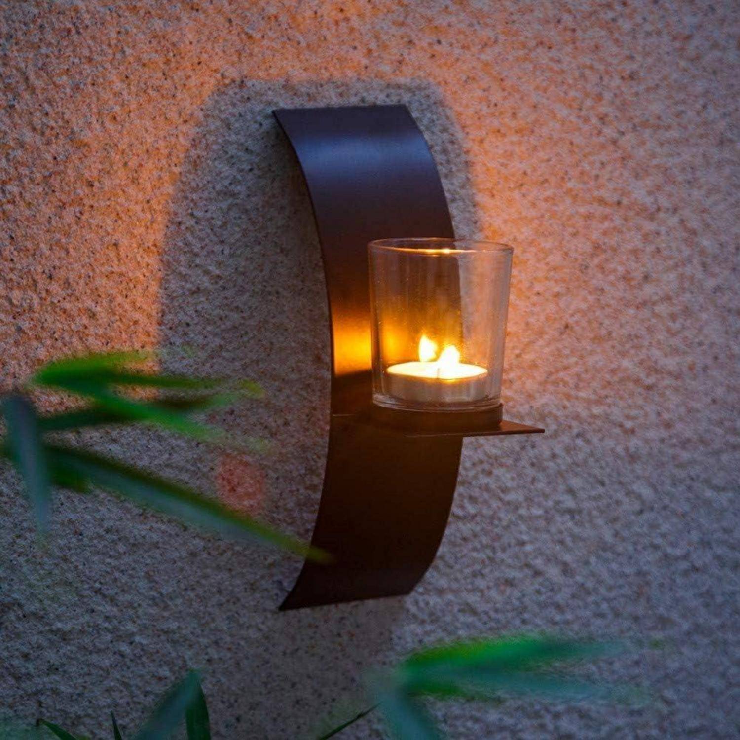 Eine minimalistische Wandleuchte mit einer brennenden Kerze beleuchtet eine strukturierte beige Wand, umgeben von grünen Blättern im Vordergrund.