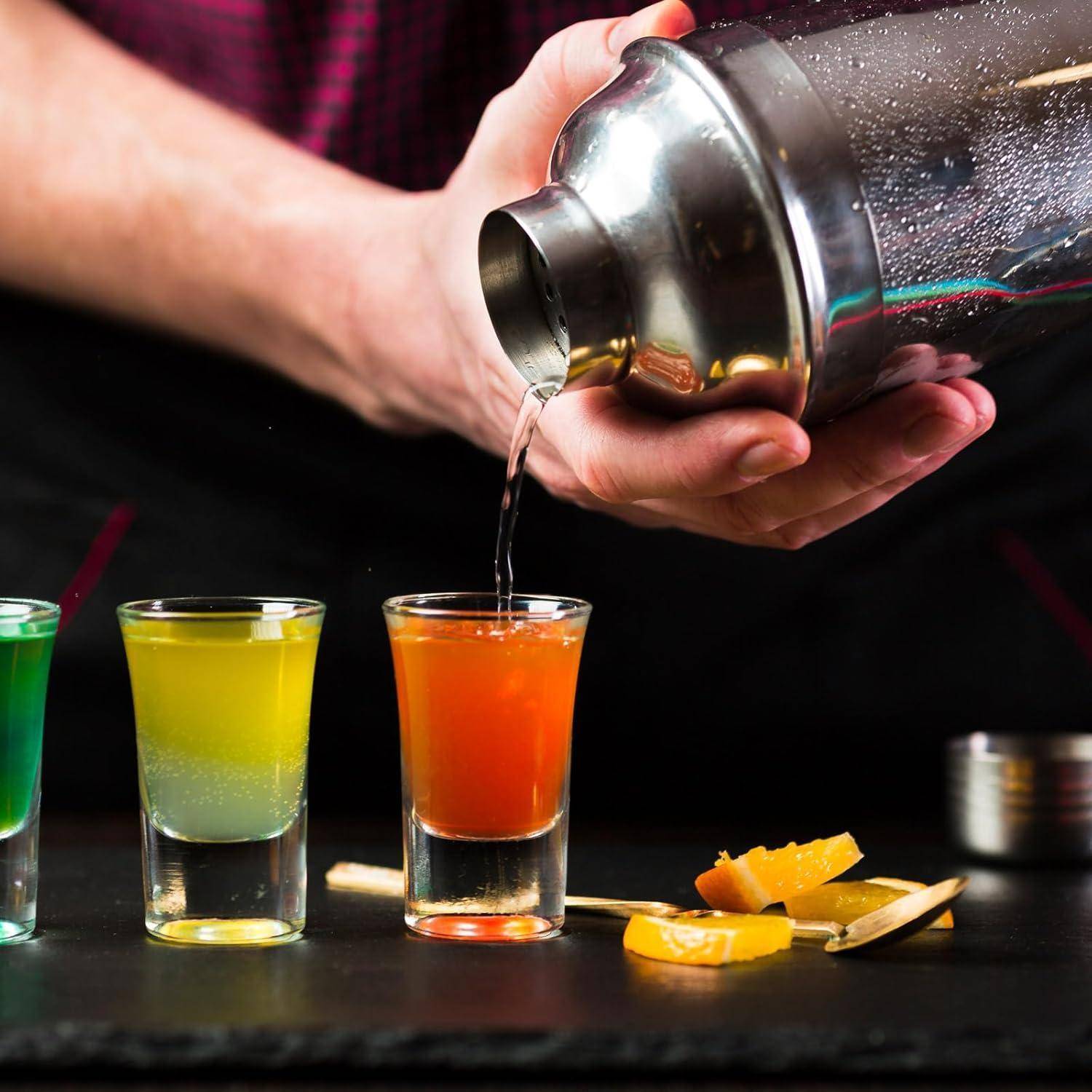Ein Barkeeper gießt einen bunten Cocktail aus einem Shaker in ein Glas auf einer Bartheke, wobei zwei weitere gefüllte Gläser und Orangengarnierungen in der Nähe stehen.