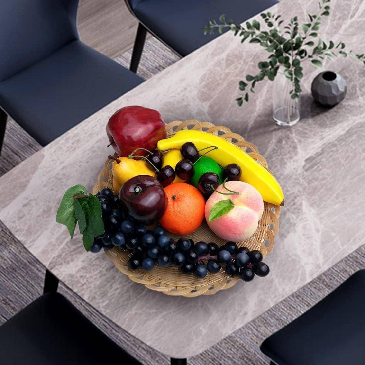 Ein Korb mit verschiedenen Früchten, darunter ein Apfel, eine Banane, Orangen und Trauben, steht auf einem Esstisch mit Stühlen darum herum.
