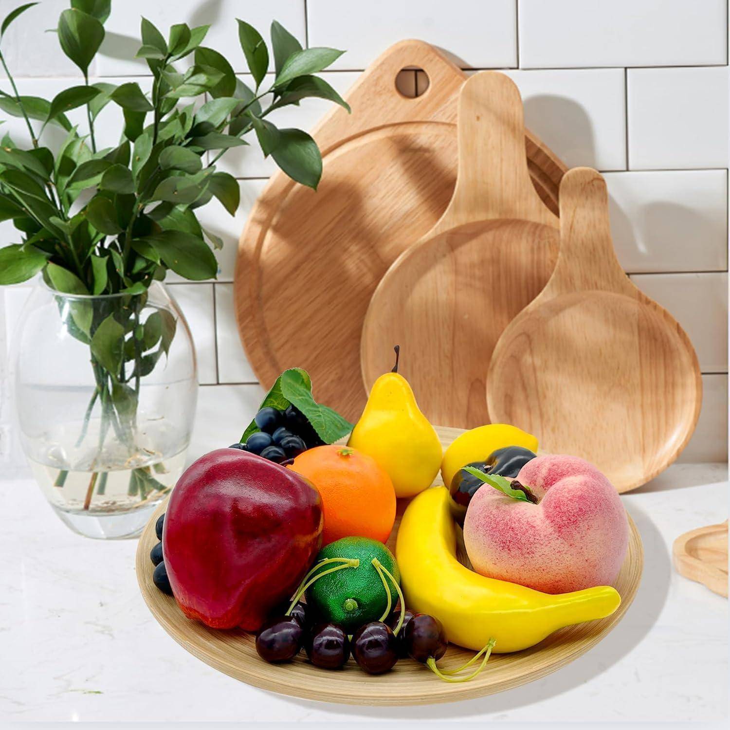 Eine Platte mit verschiedenen künstlichen Früchten, darunter ein roter Apfel, eine Banane und ein Pfirsich, wird auf einer Küchenablage mit Holzschneidebrettern und einer Vase mit grünen Blättern im Hintergrund präsentiert.